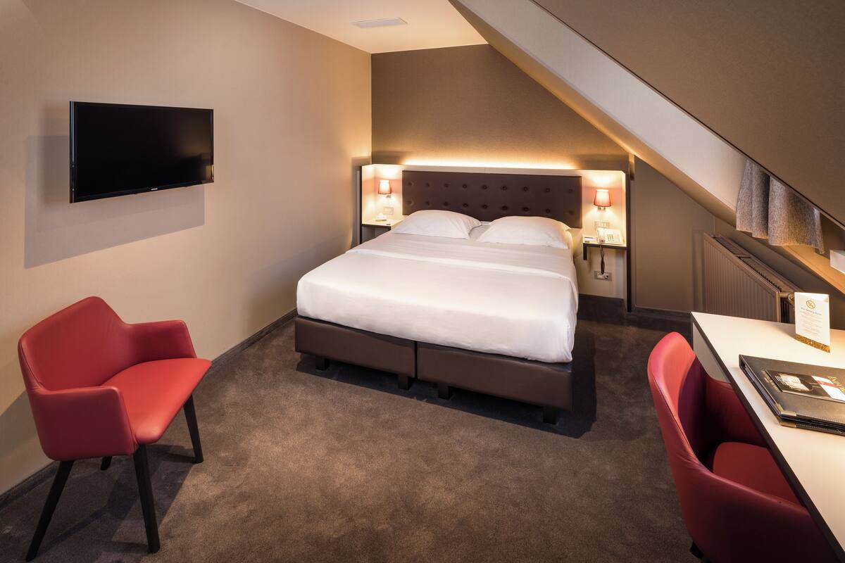 Hotellihuone, jossa on sänky, seinälle asennettu televisio, työpöytä tuolilla ja portaikko.