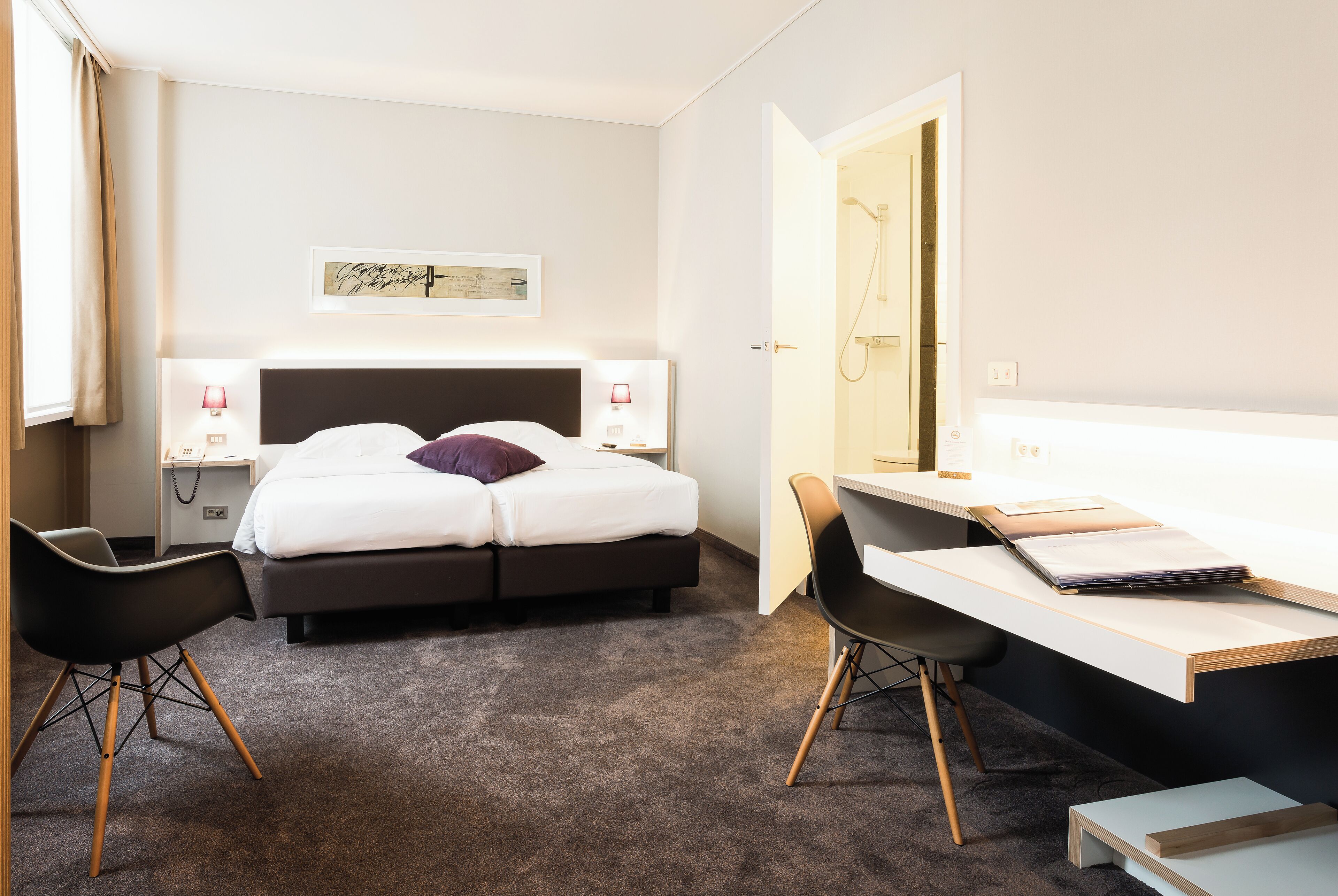 A hotel room with a bed, two bedside tables with lamps, a desk with a chair, and a shower area.