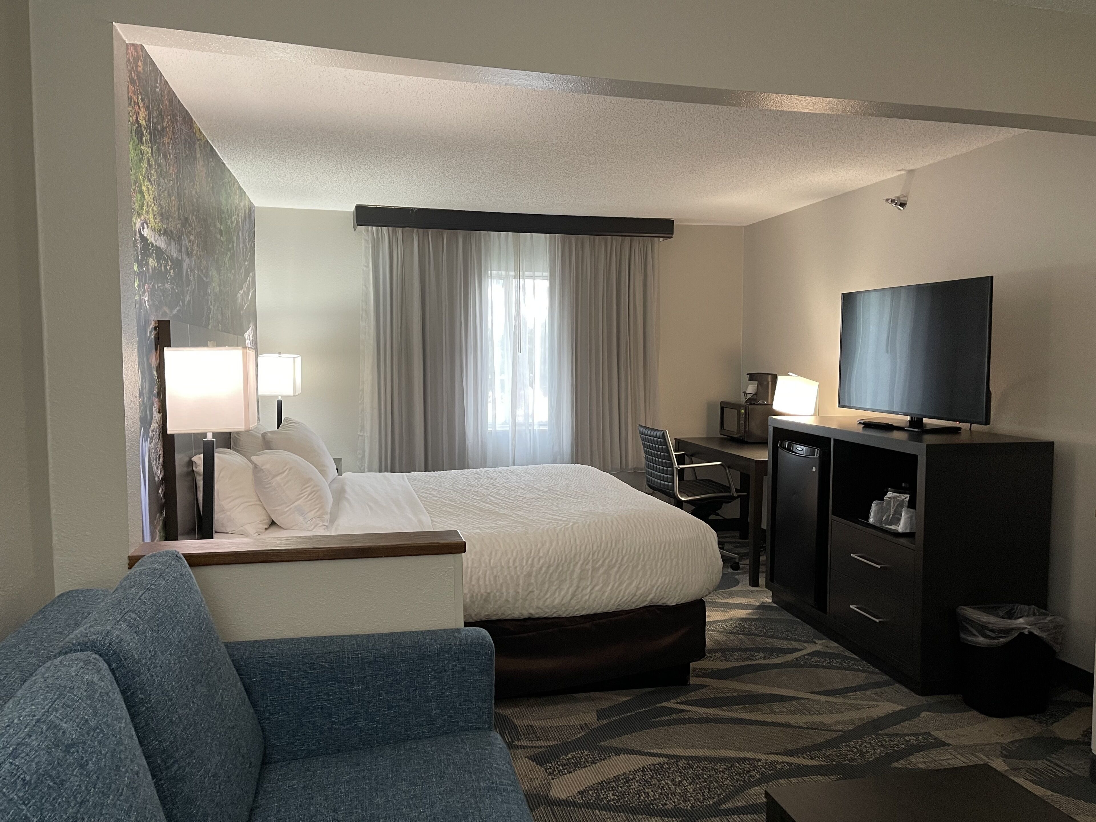 Una habitación de hotel con una cama, un escritorio con televisor, una silla, una lámpara y un ventanal con cortinas.