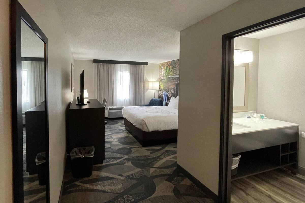 A hotel room with a bed, a desk, a dresser, a bathroom with a sink and mirror, and a patterned carpet.