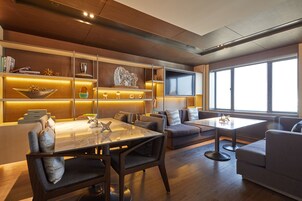 A modern living room with a dining area, wooden flooring, and large windows providing natural light.