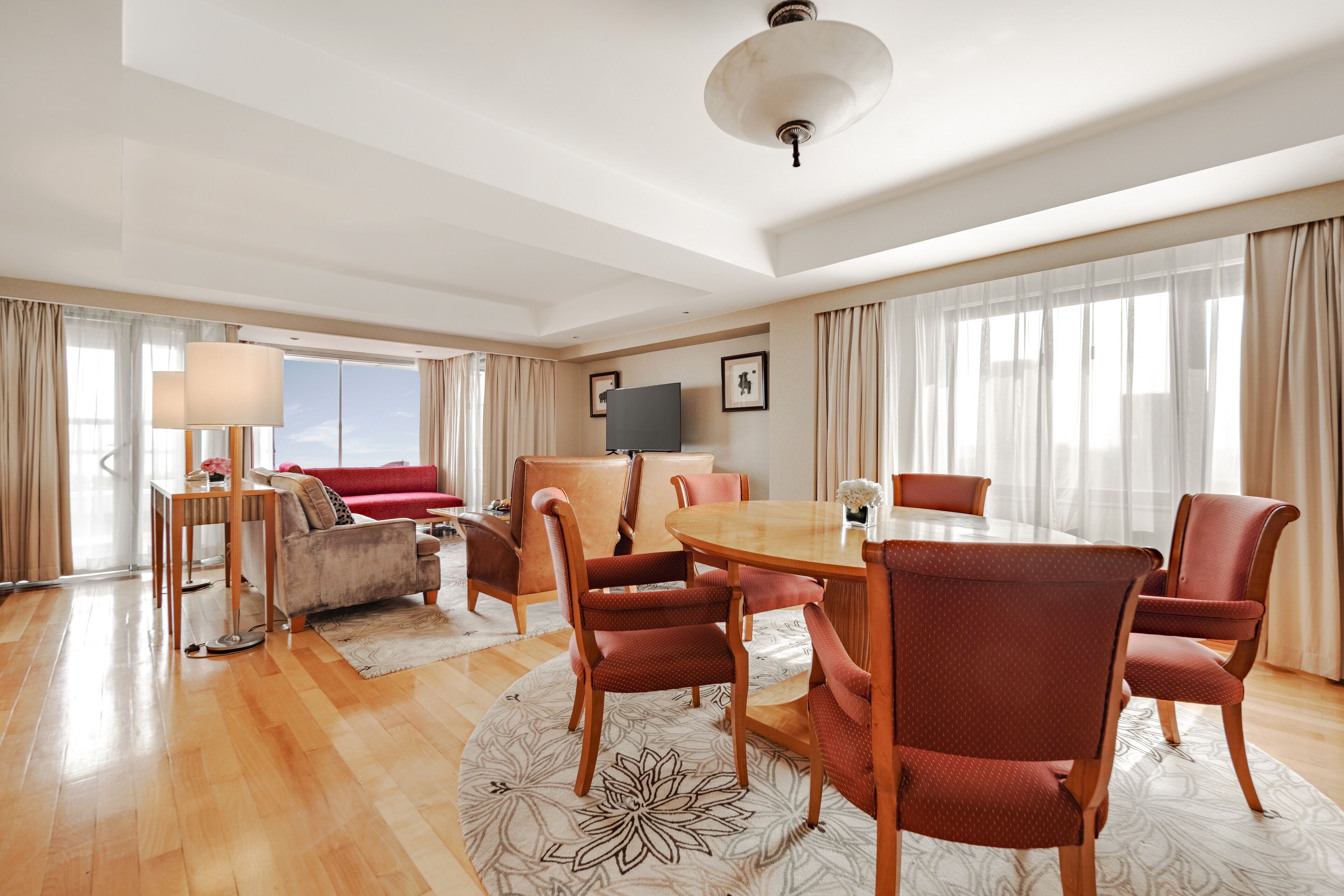 A spacious living room with a round wooden table, red upholstered chairs, a sofa, and a television on the wall.