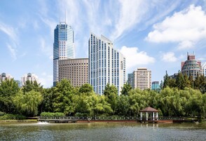 A modern hotel with a glass facade, surrounded by lush greenery and a calm lake.