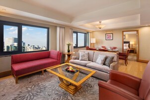 A modern hotel room with a red sofa, a grey sofa, and a wooden coffee table with fruit. Large windows offer a city view.