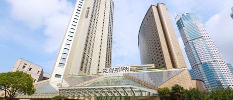 A modern hotel with a glass facade, a prominent Radisson Collection sign, and a surrounding urban landscape with other high-rise buildings.