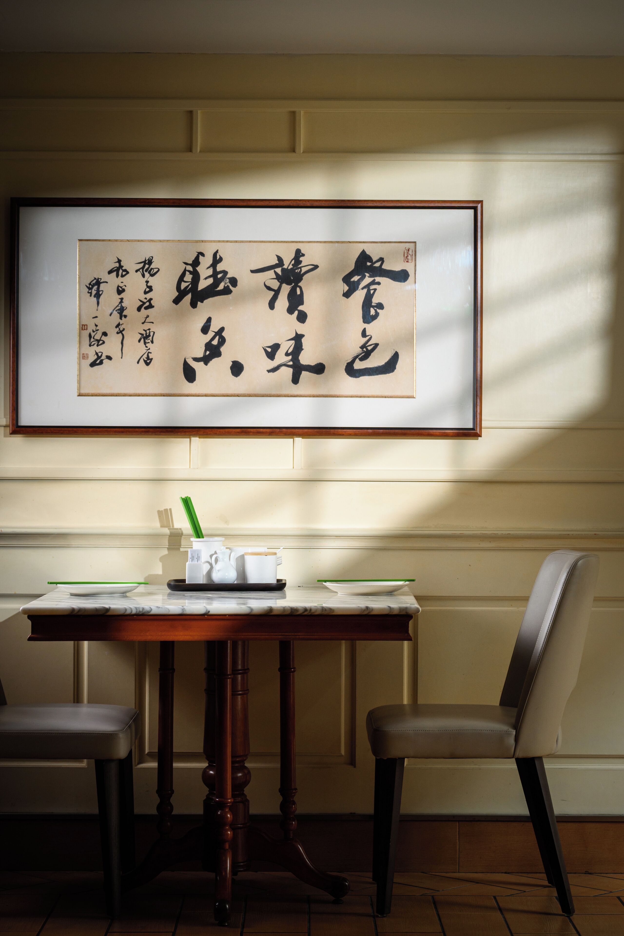 Una obra de caligrafía enmarcada en la pared, una mesa con palillos, un hervidor de té y una taza, y dos sillas en una habitación con suelo de madera.