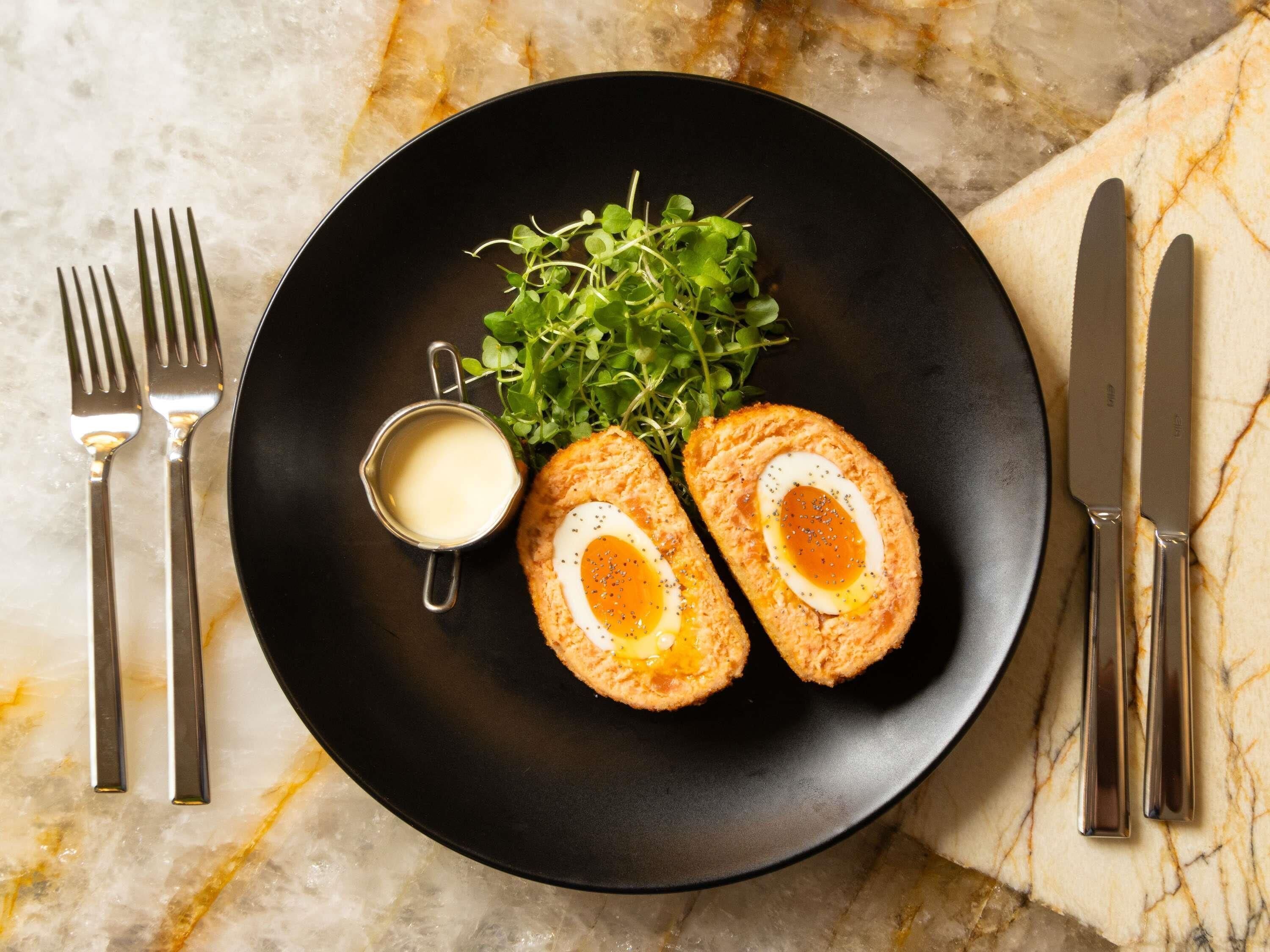 A black plate with two halves of a Scotch egg, a small bowl of sauce, and some greens on a marble surface.