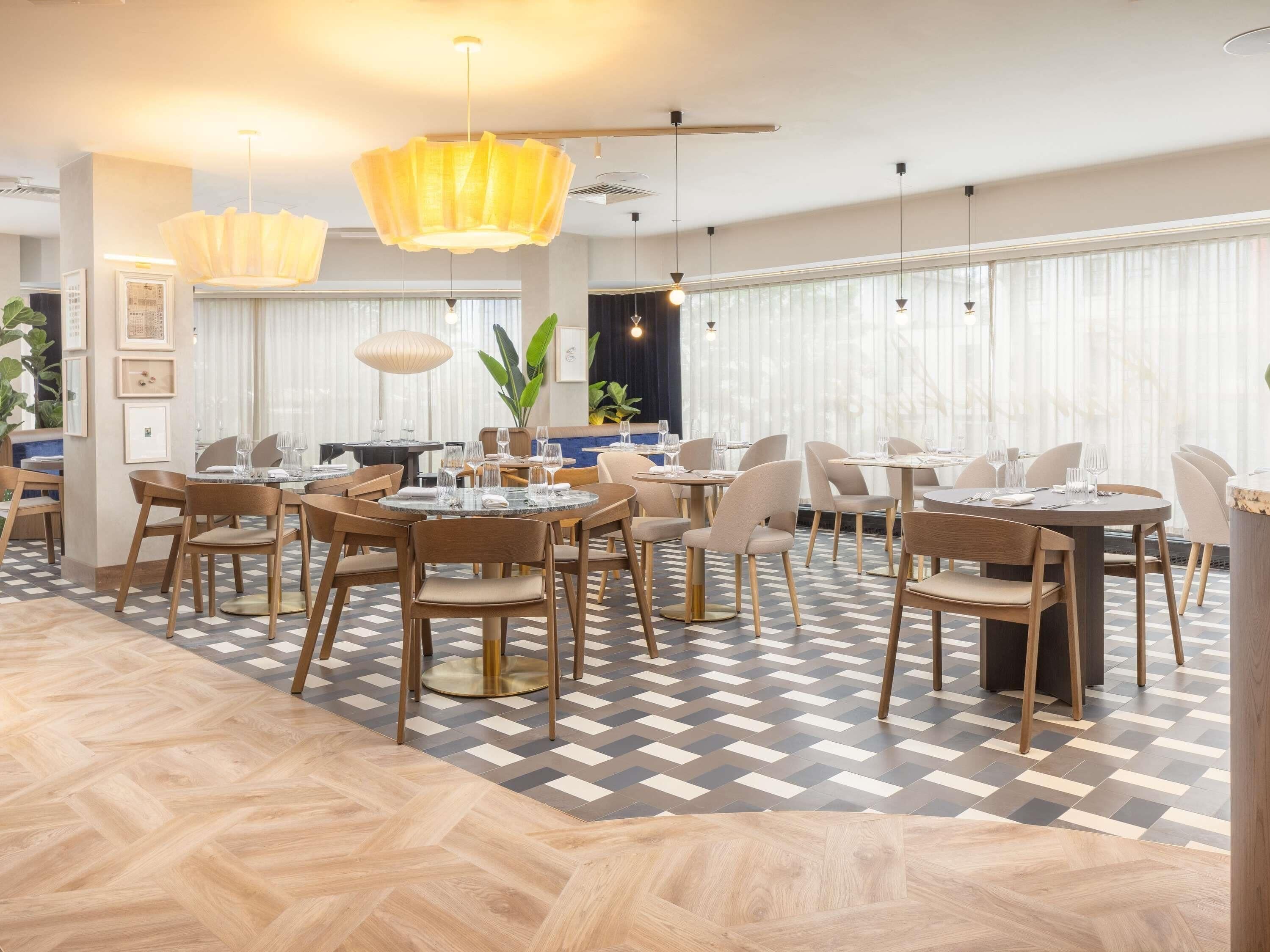A modern dining area with round tables and chairs, large windows with sheer curtains, and a herringbone patterned floor.