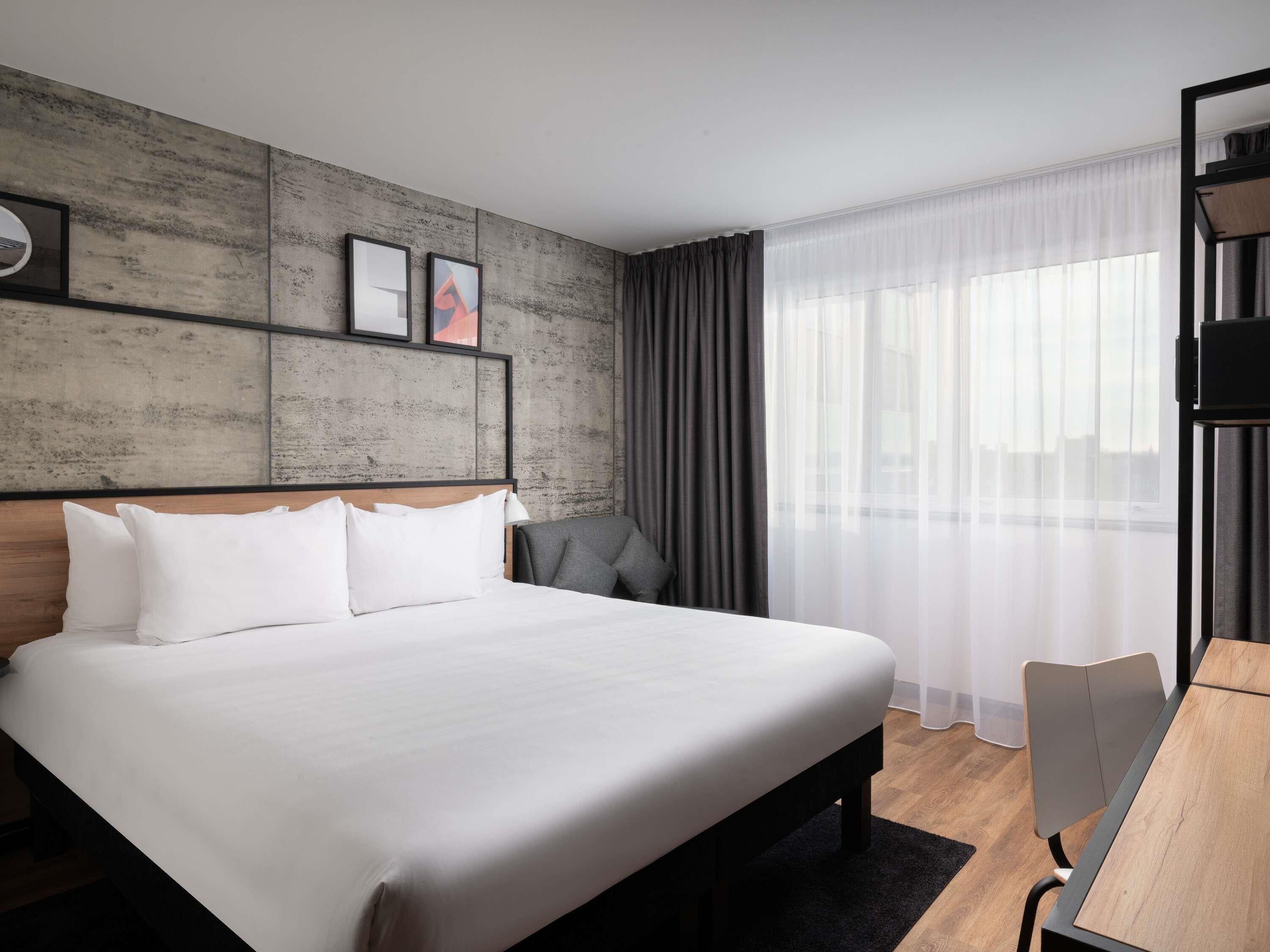 A modern hotel room with a large bed, a desk, and a view of the city through sheer curtains.