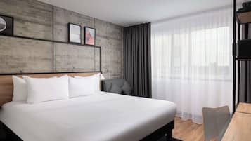 A modern hotel room with a large bed, a desk, and a view of the city through sheer curtains.
