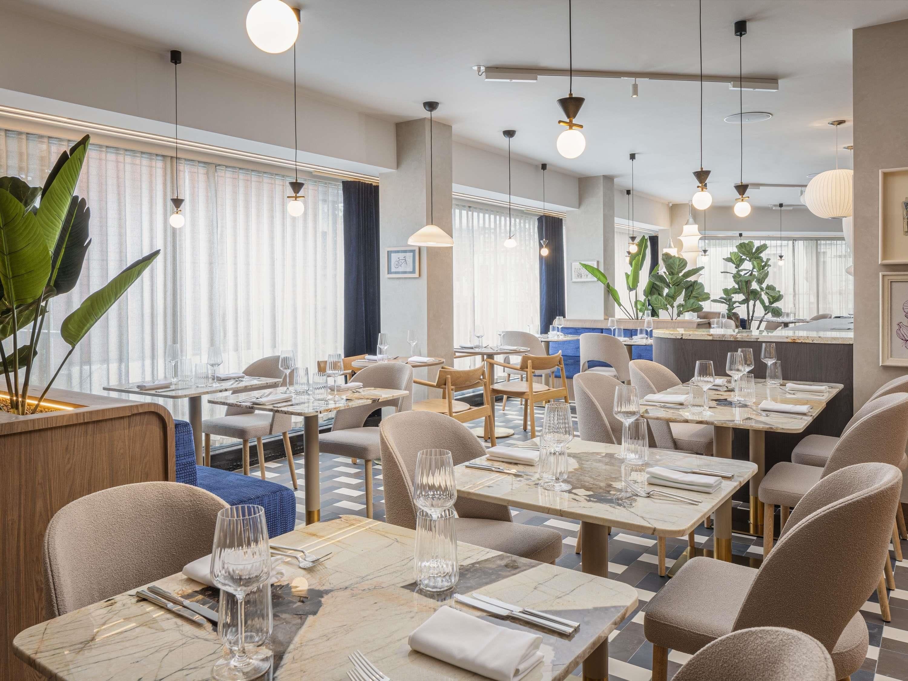 A modern dining area with multiple tables and chairs, large windows with curtains, and hanging pendant lights.