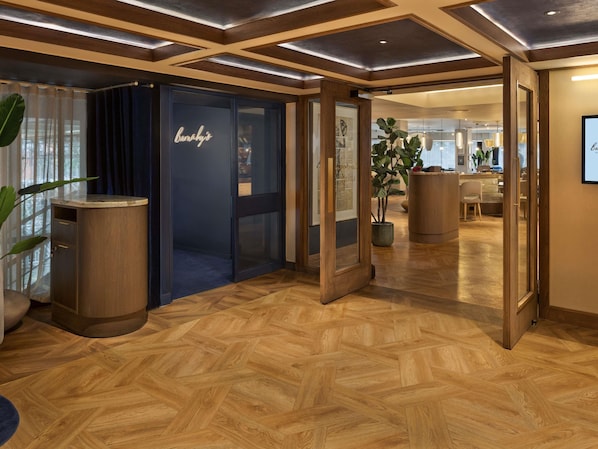 Un vestíbulo de hotel con piso de madera en patrón de espiga, un mostrador de recepción y una puerta de cristal con un cartel.
