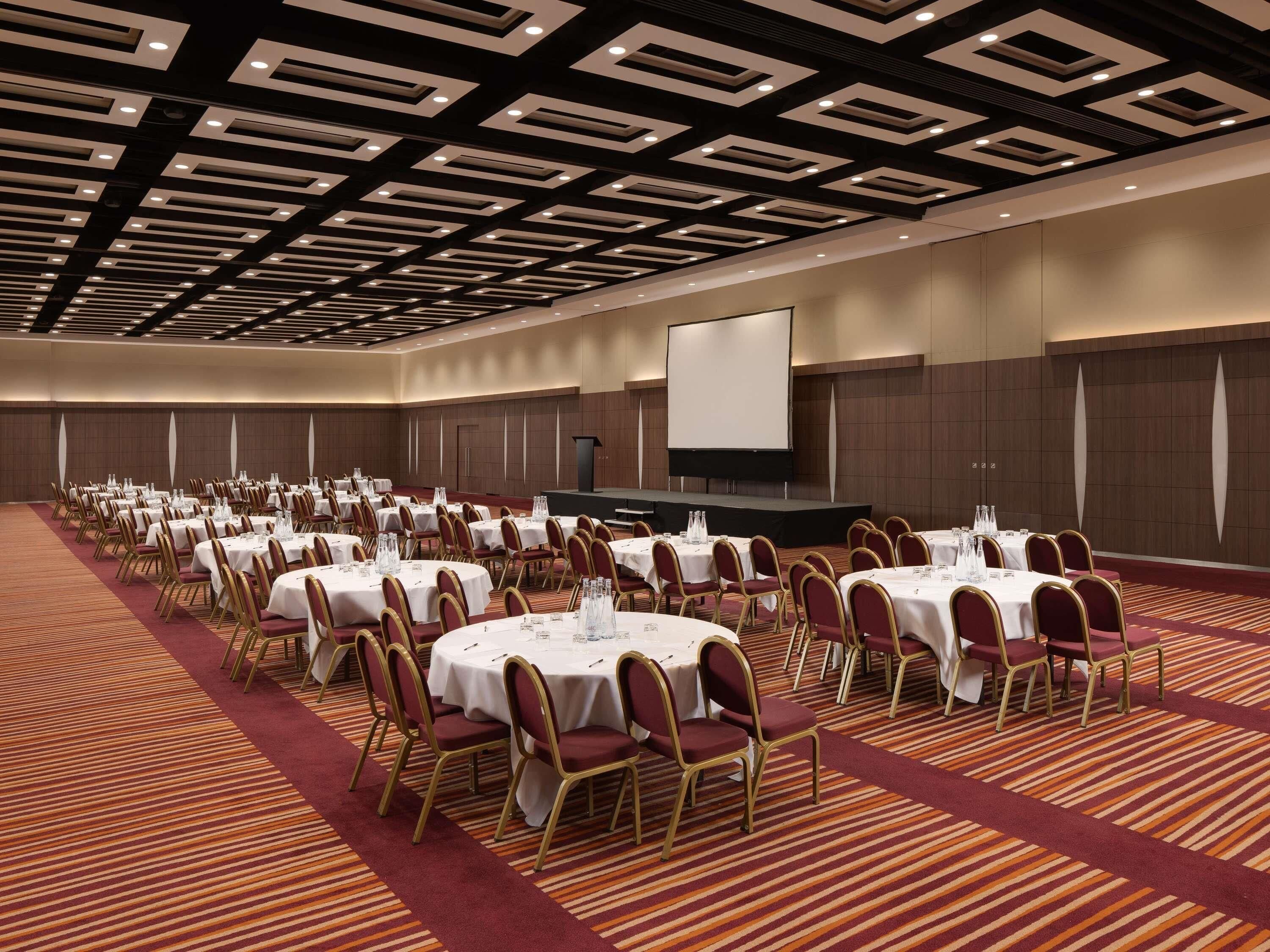 A large conference room with multiple round tables, chairs, and a stage with a screen.