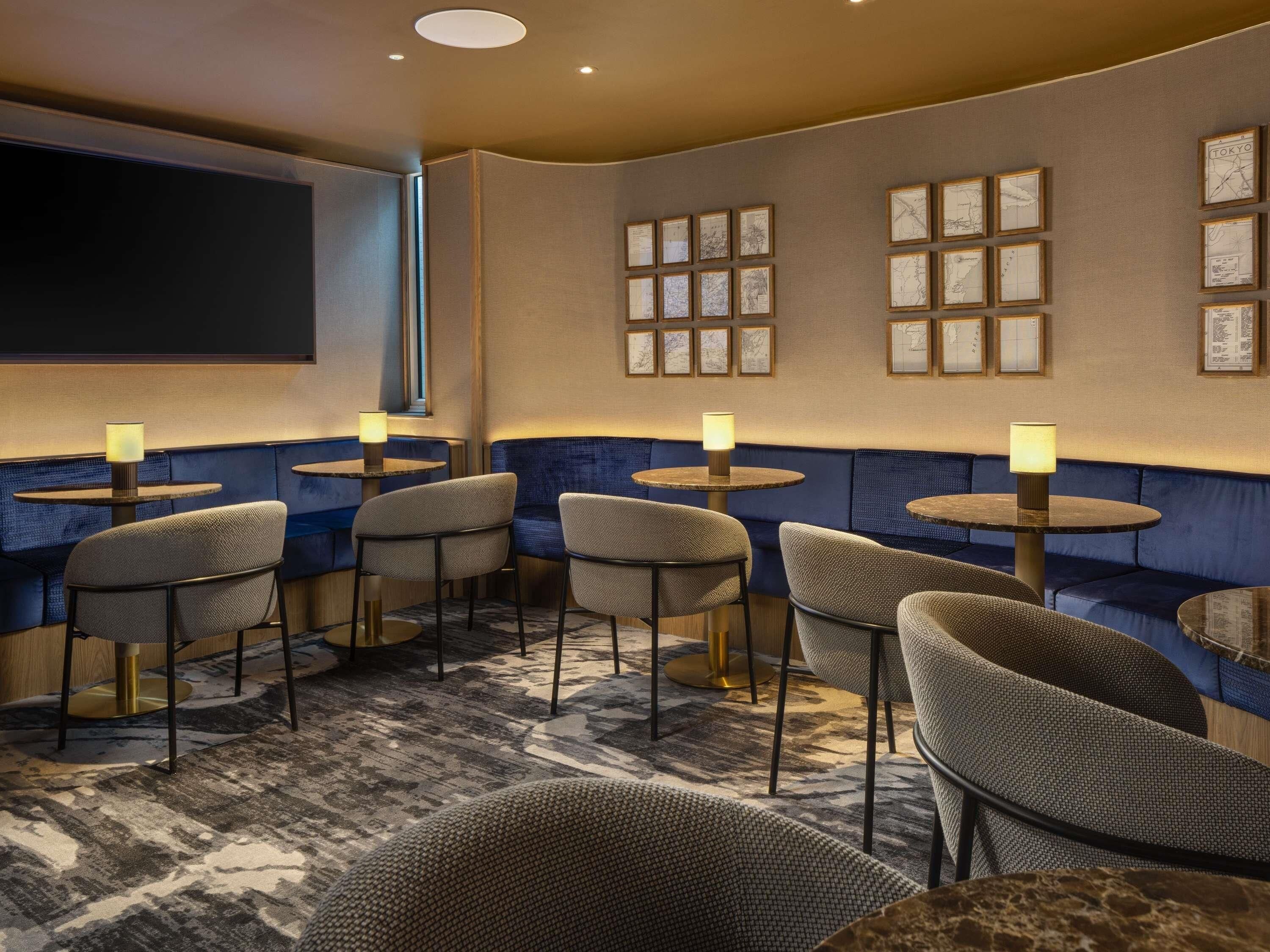 A hotel room with a seating area featuring round tables and chairs, a large flat-screen TV mounted on the wall, and framed maps on the wall.