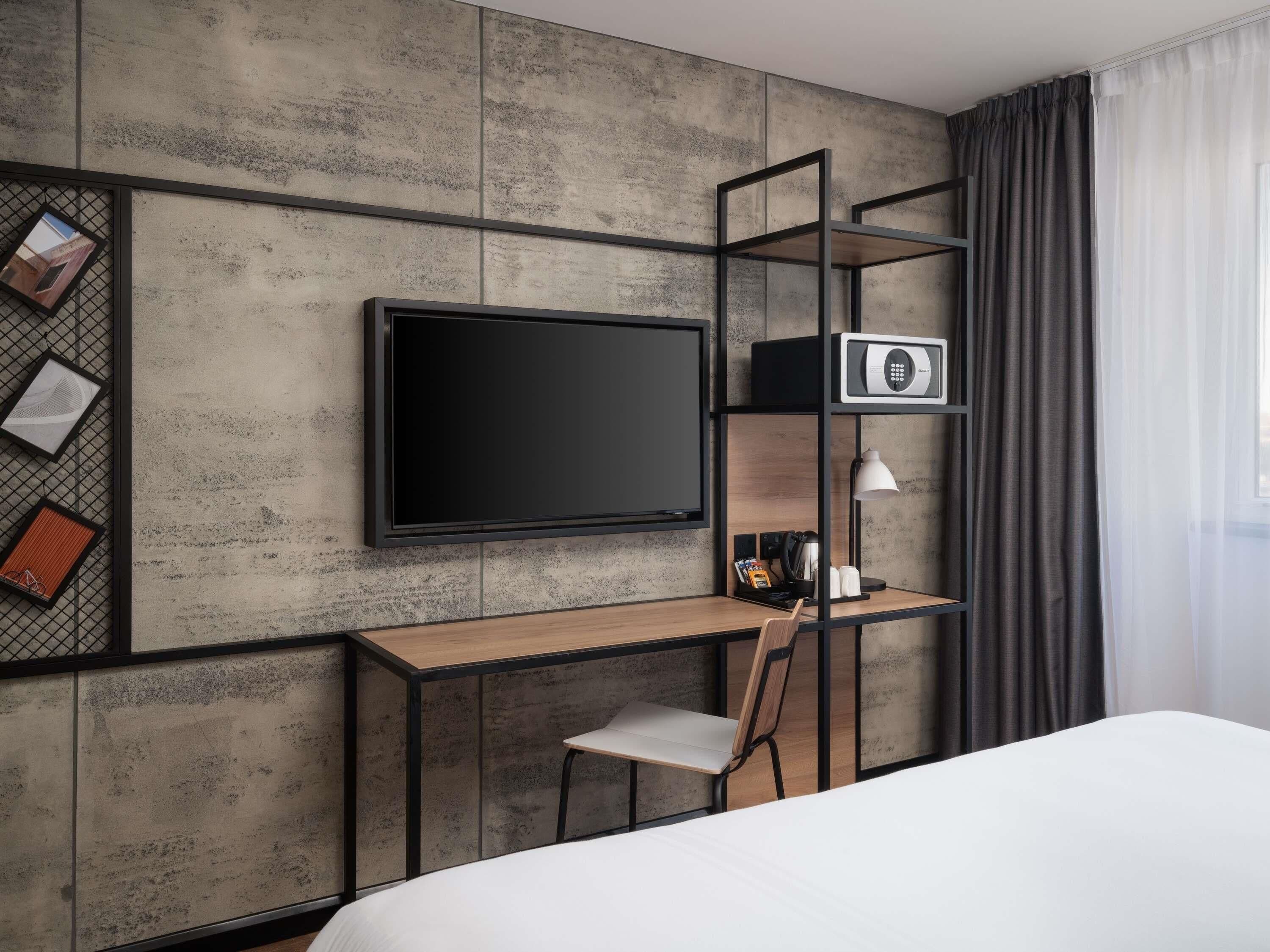 A modern hotel room with a large flat-screen TV mounted on a textured wall, a wooden desk with a chair, a small shelf with a lamp, and a bed with white bedding.