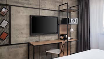 A modern hotel room with a large flat-screen TV mounted on a textured wall, a wooden desk with a chair, a small shelf with a lamp, and a bed with white bedding.