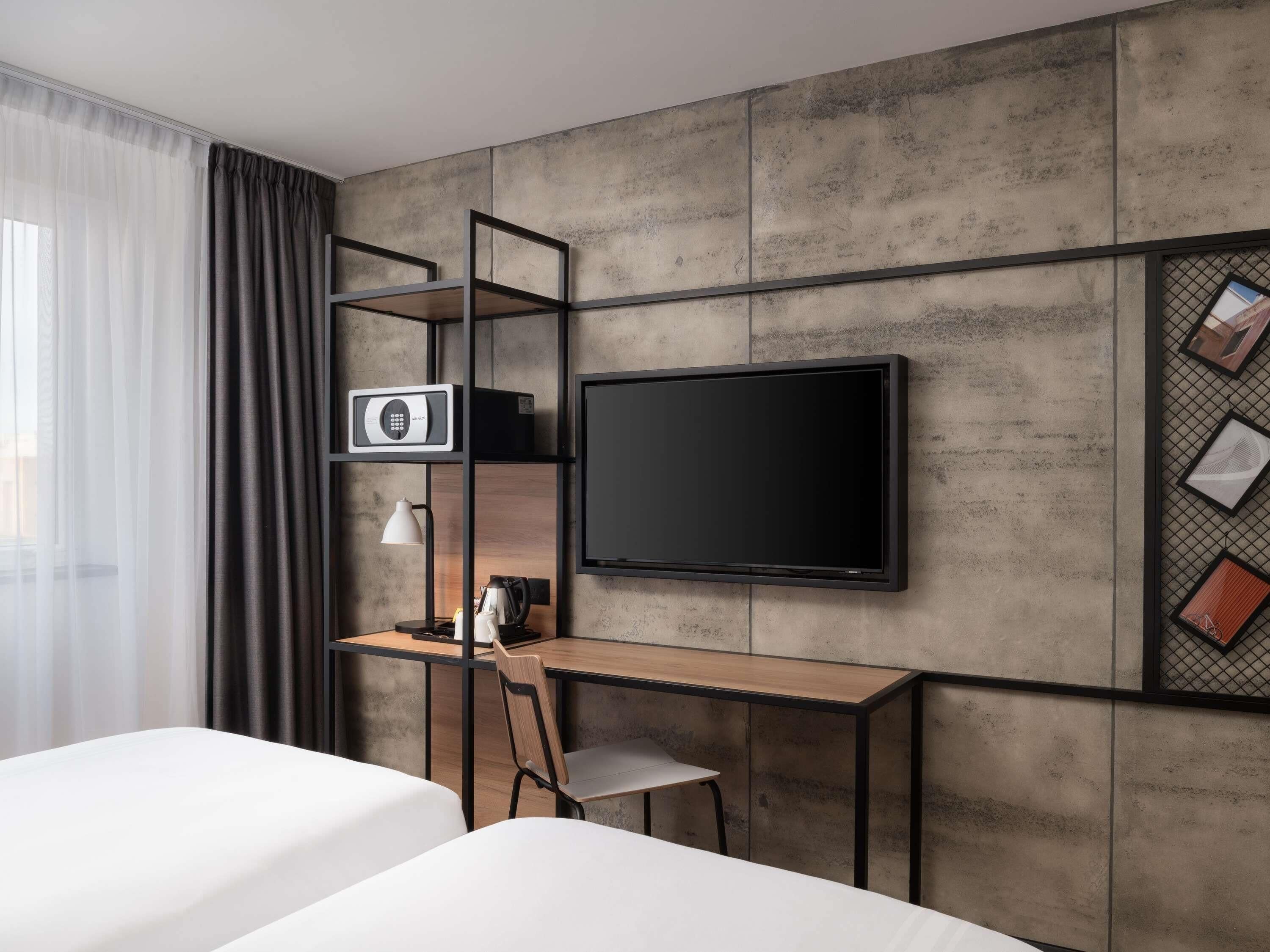 Habitación de hotel con cama, escritorio con silla, televisor, máquina de café y ventana con cortinas.