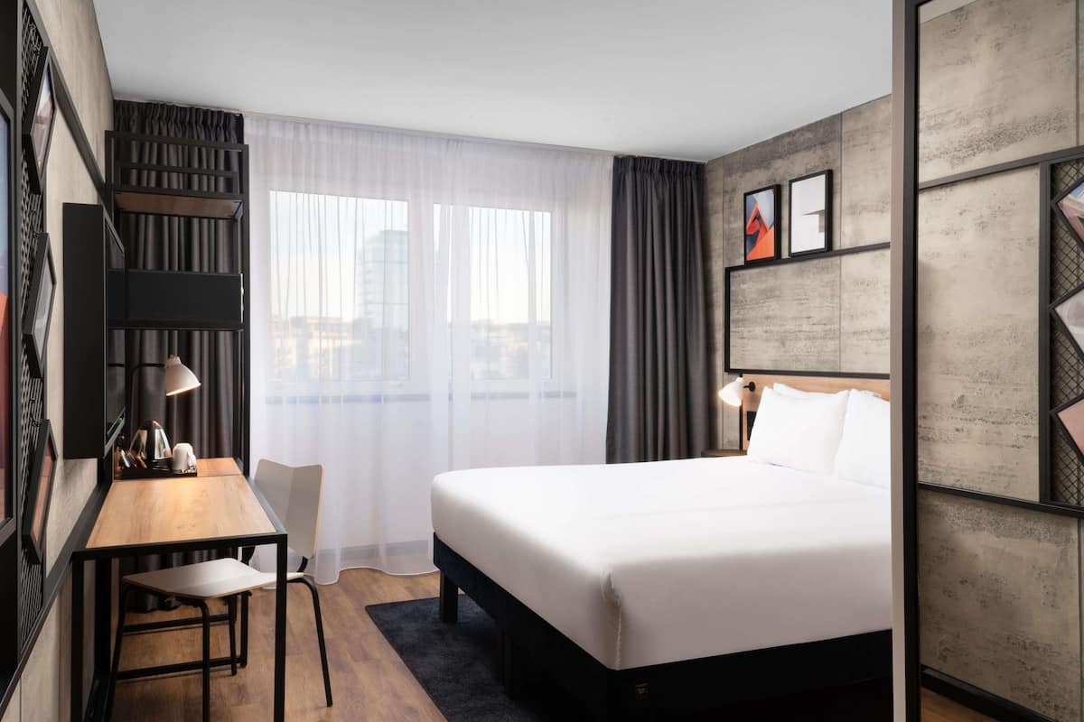 Una habitación de hotel con cama, escritorio, silla y un amplio ventanal con cortinas.