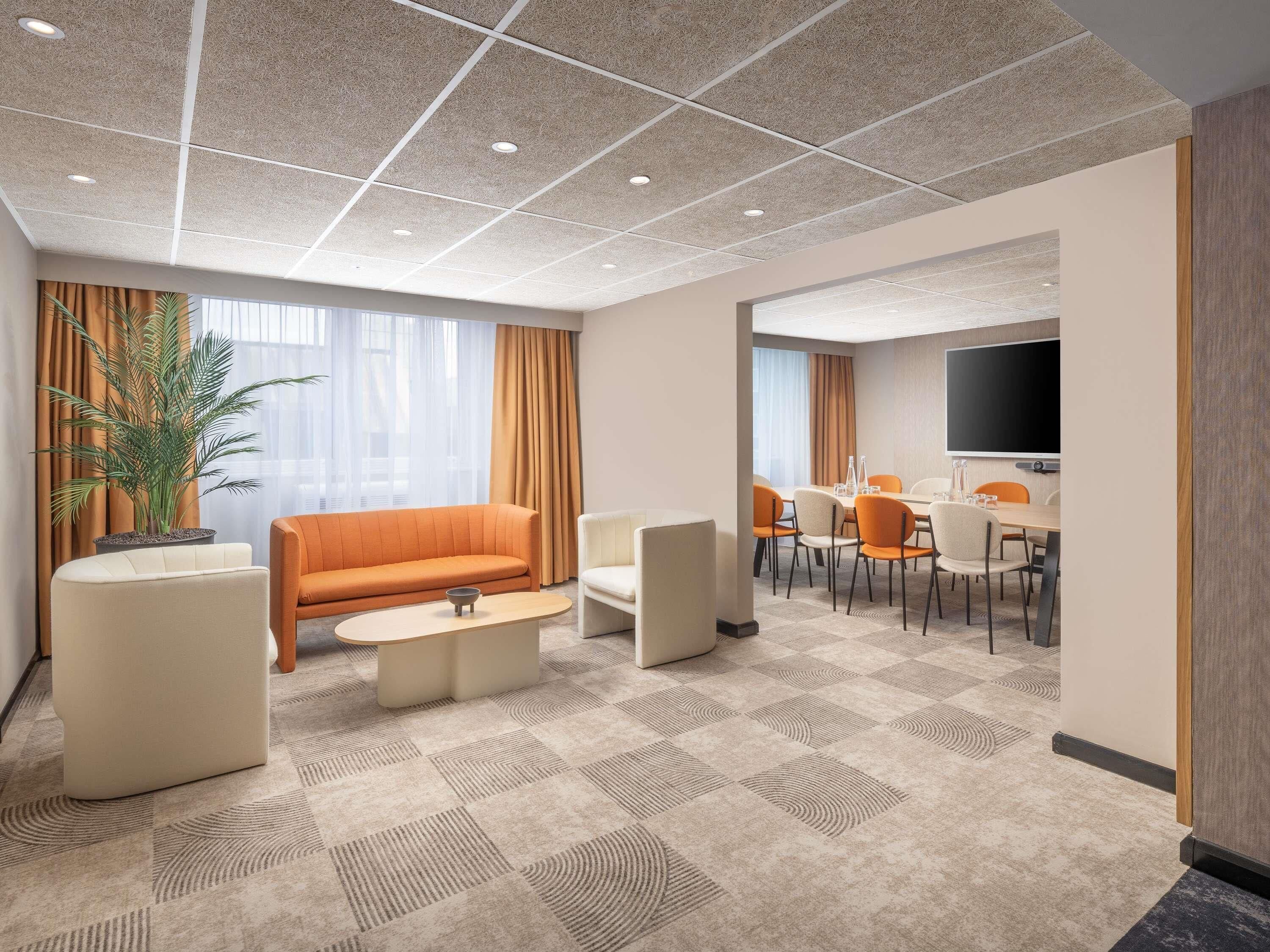 A hotel room with a sofa, a coffee table, a dining table with chairs, a television, and a potted plant.