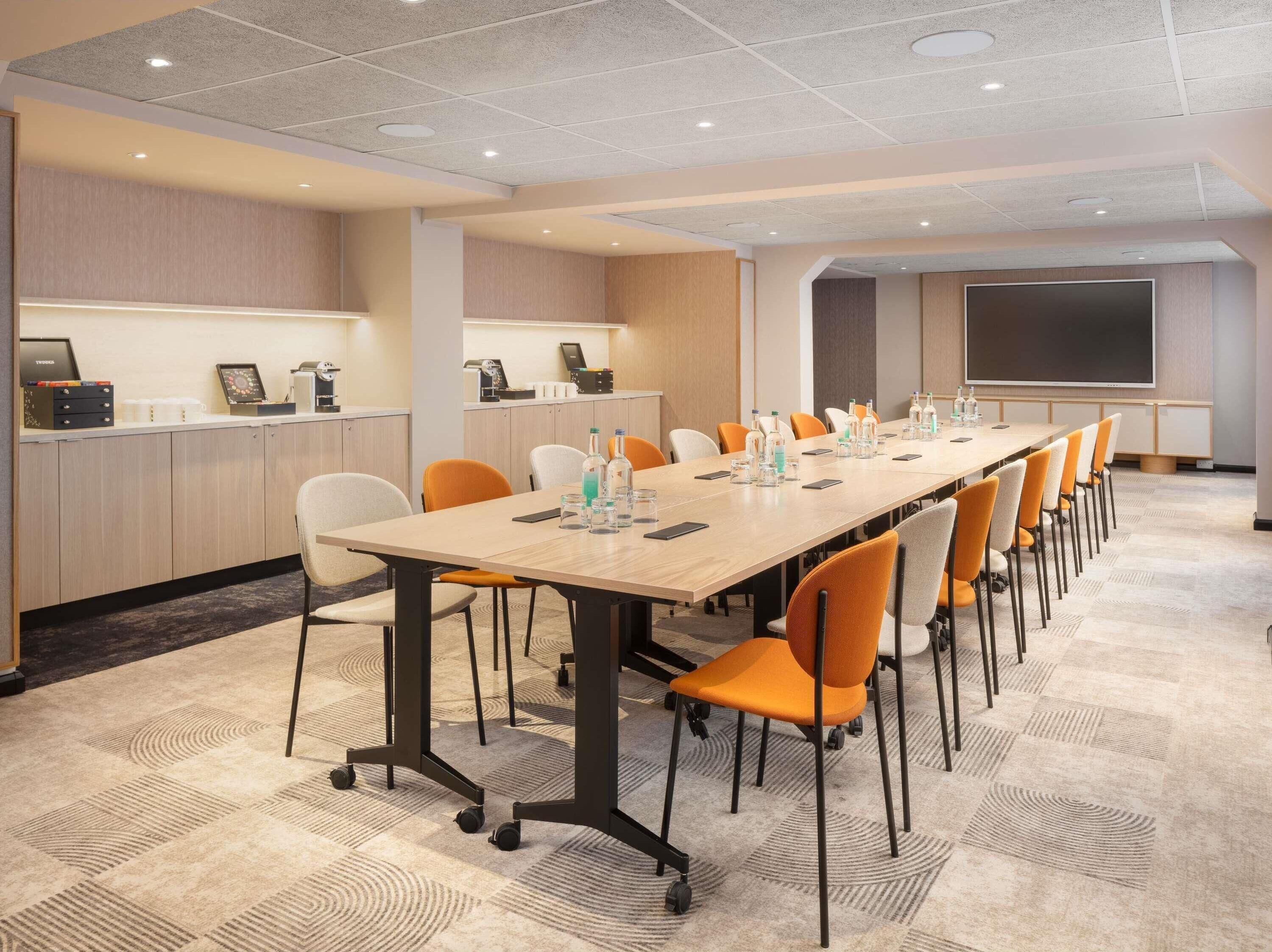 Una sala de conferencias con una larga mesa, sillas, una pantalla de proyector y una zona de kitchenette.