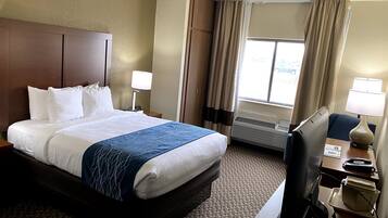 A hotel room with a large bed, two bedside tables with lamps, a desk with a computer, and a window with curtains.