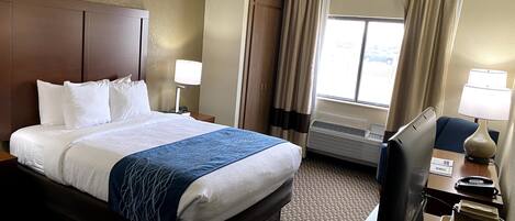 A hotel room with a large bed, two bedside tables with lamps, a desk with a computer, and a window with curtains.