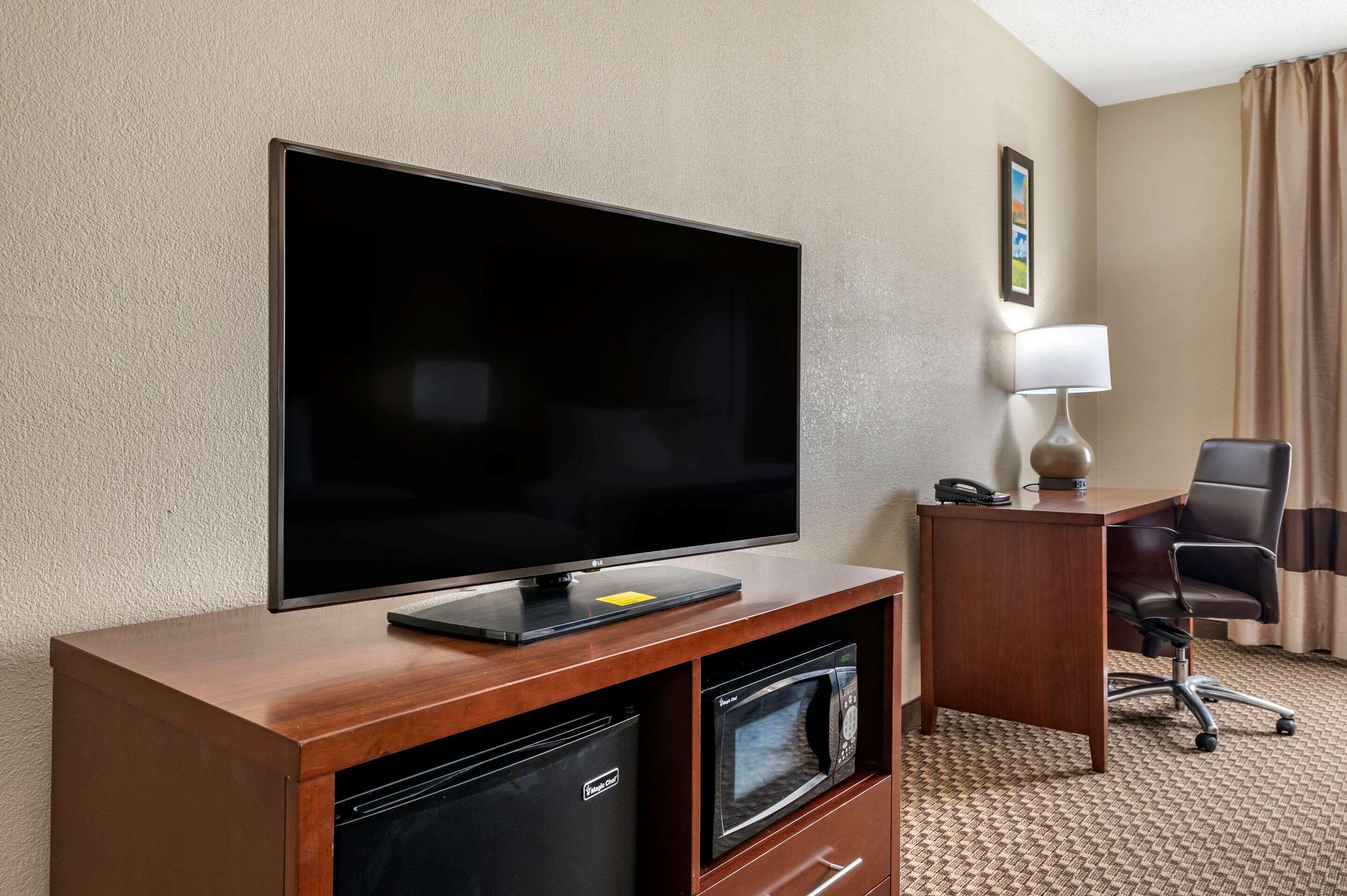 Habitación de hotel con una televisión de pantalla plana sobre un soporte de madera, un escritorio con una silla, una lámpara y un minibar.