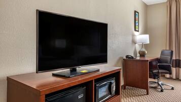 Habitación de hotel con una televisión de pantalla plana sobre un soporte de madera, un escritorio con una silla, una lámpara y un minibar.