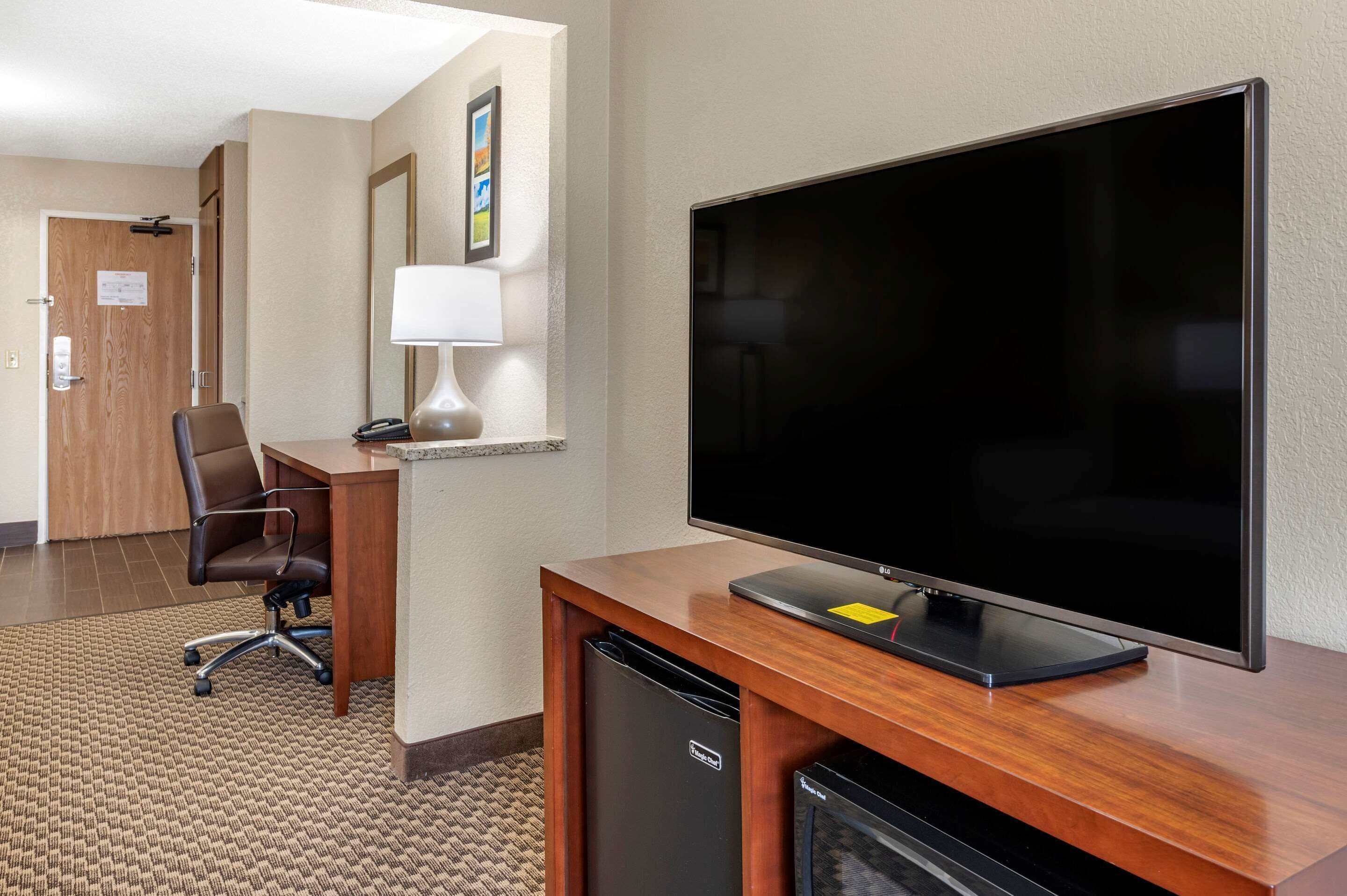 Habitación de hotel con un escritorio de madera, un televisor de pantalla plana, una silla, una lámpara y un refrigerador pequeño.