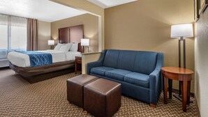 A hotel room with a bed, a blue sofa, a wooden side table, and a lamp. - Comfort Inn Near Kokomo Speedway (Kokomo)