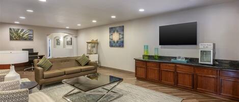 A modern living room with a sofa, armchairs, a glass coffee table, and a kitchen area with wooden cabinets and a countertop.