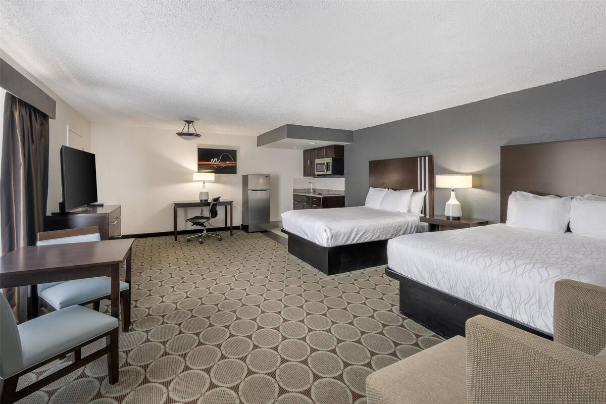 A hotel room with two beds, a desk, a chair, a television, and a kitchenette.