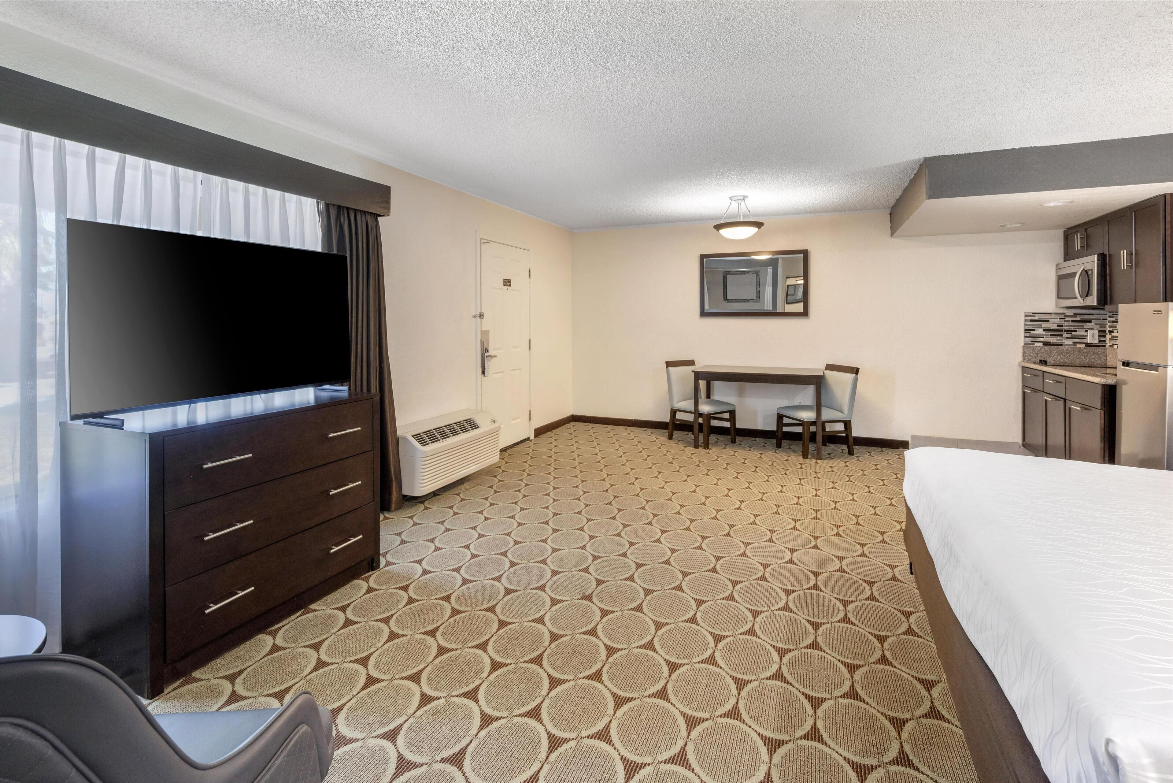 A hotel room with a bed, a TV, a desk with chairs, a kitchenette, and a patterned carpet.