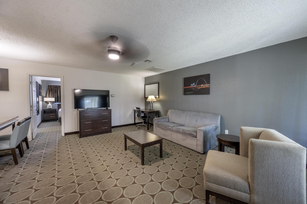 A hotel room with a sofa, armchair, TV, desk, and patterned carpet.