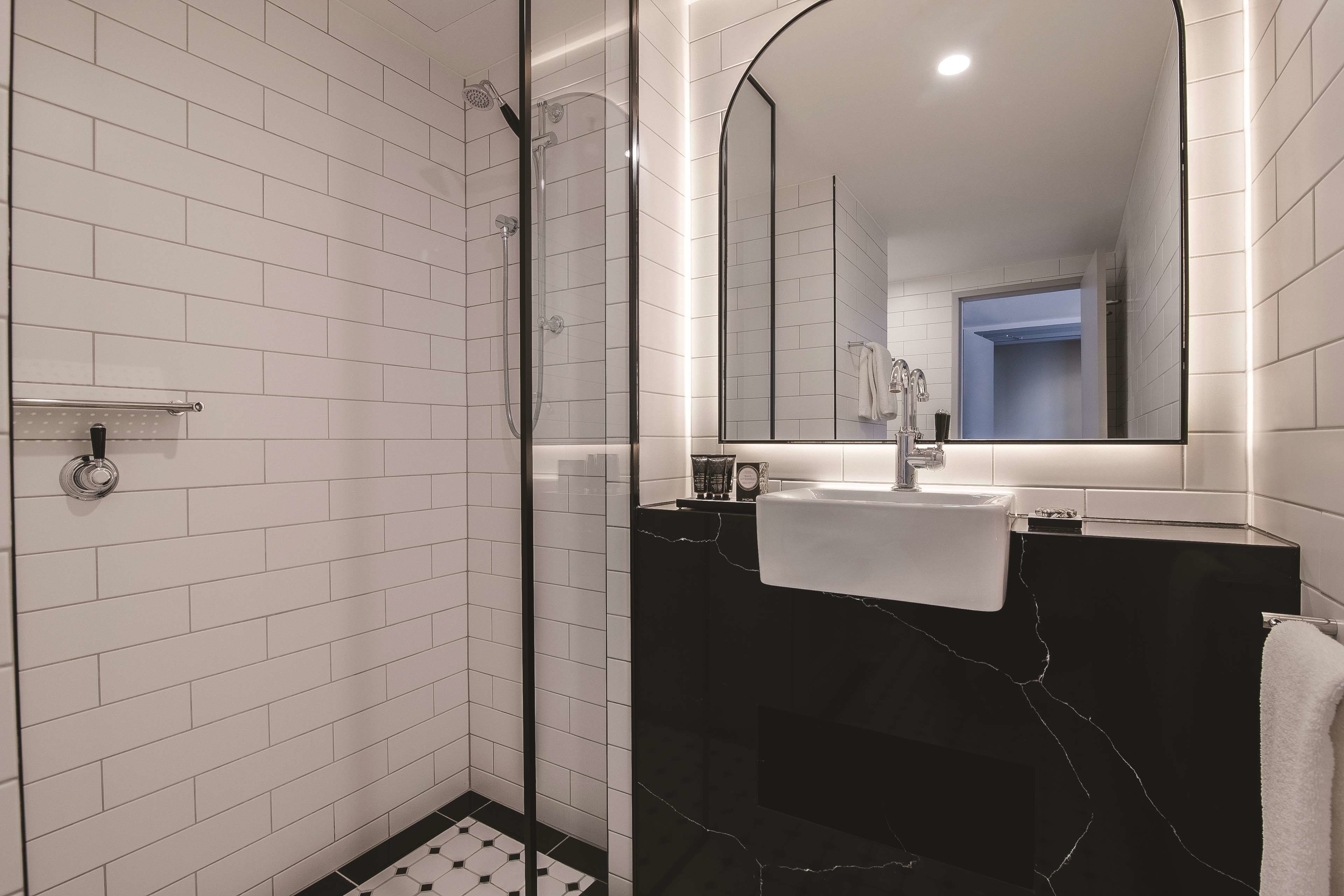 A modern bathroom with white subway tiles, a glass-enclosed shower, a black marble vanity, and a large mirror.