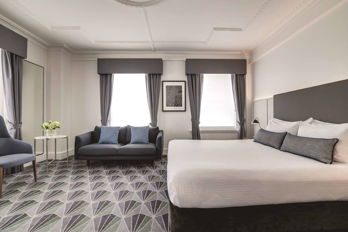 Ein Hotelzimmer mit einem großen Bett, einer Couch, einem kleinen Tisch mit Blumenvase und zwei mit Vorhängen versehenen Fenstern.