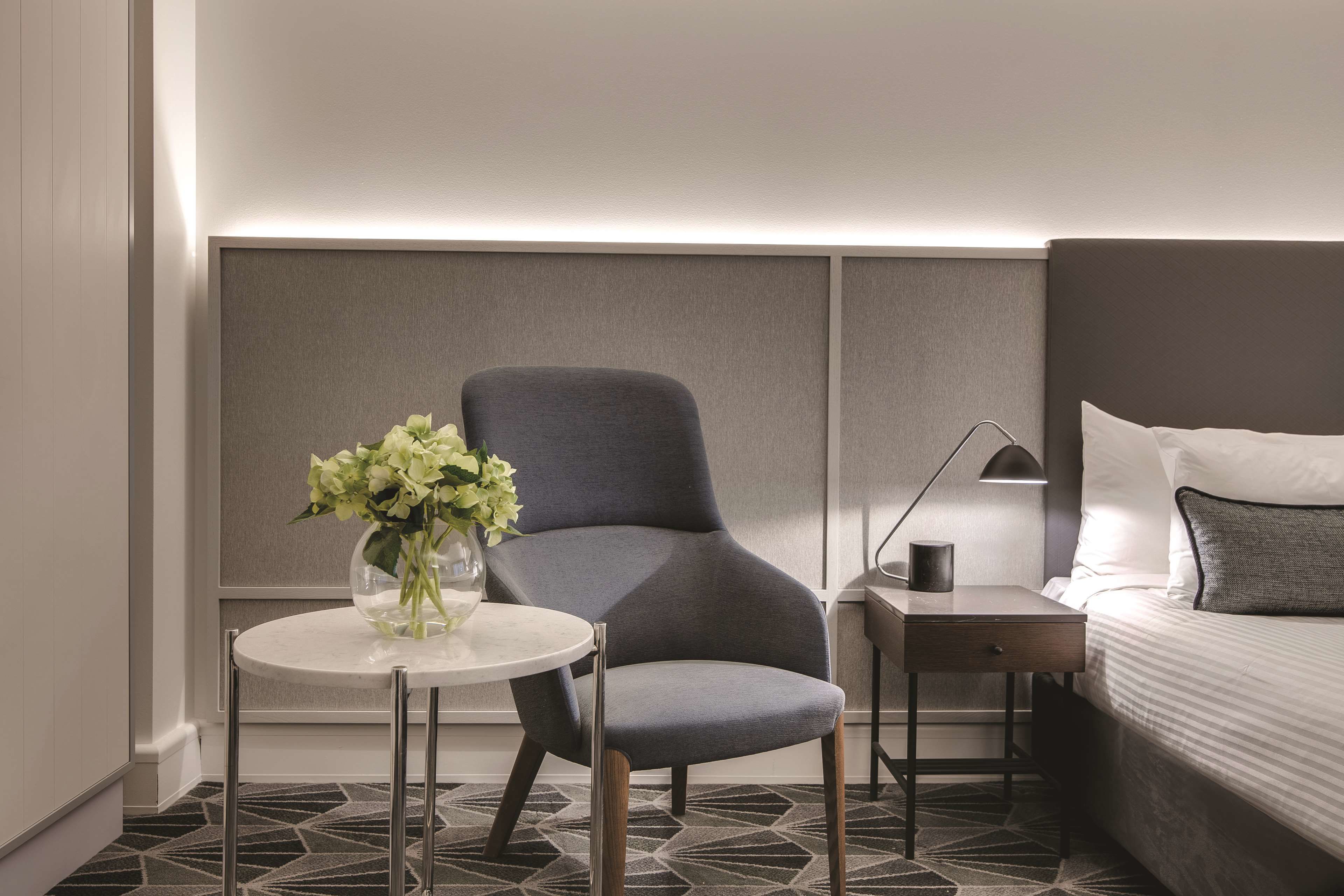 Una habitación de hotel moderna con un sillón tapizado en gris, una mesita redonda con un florero, una mesita de noche con lámpara, y una cama bien hecha.