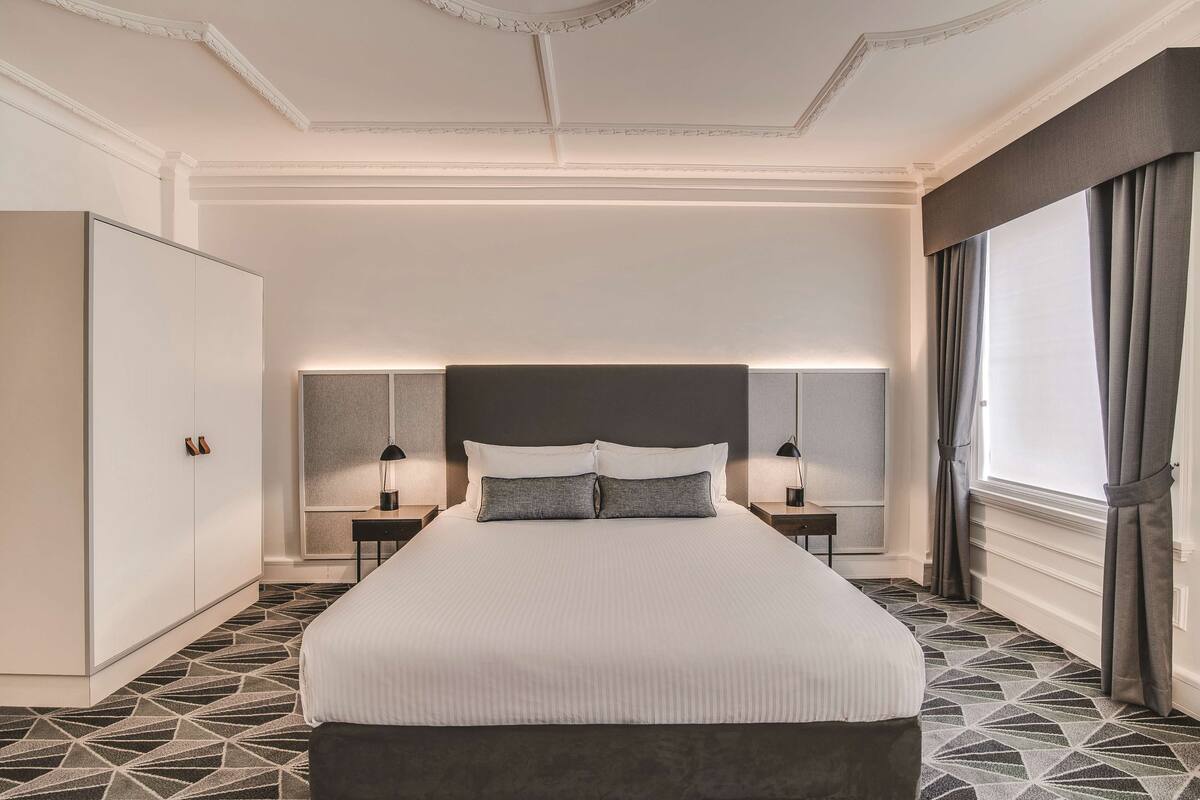 Ein ordentlich eingerichtetes Hotelzimmer mit einem großen Bett, Nachttischen, einem Kleiderschrank und einem Fenster mit Vorhängen.