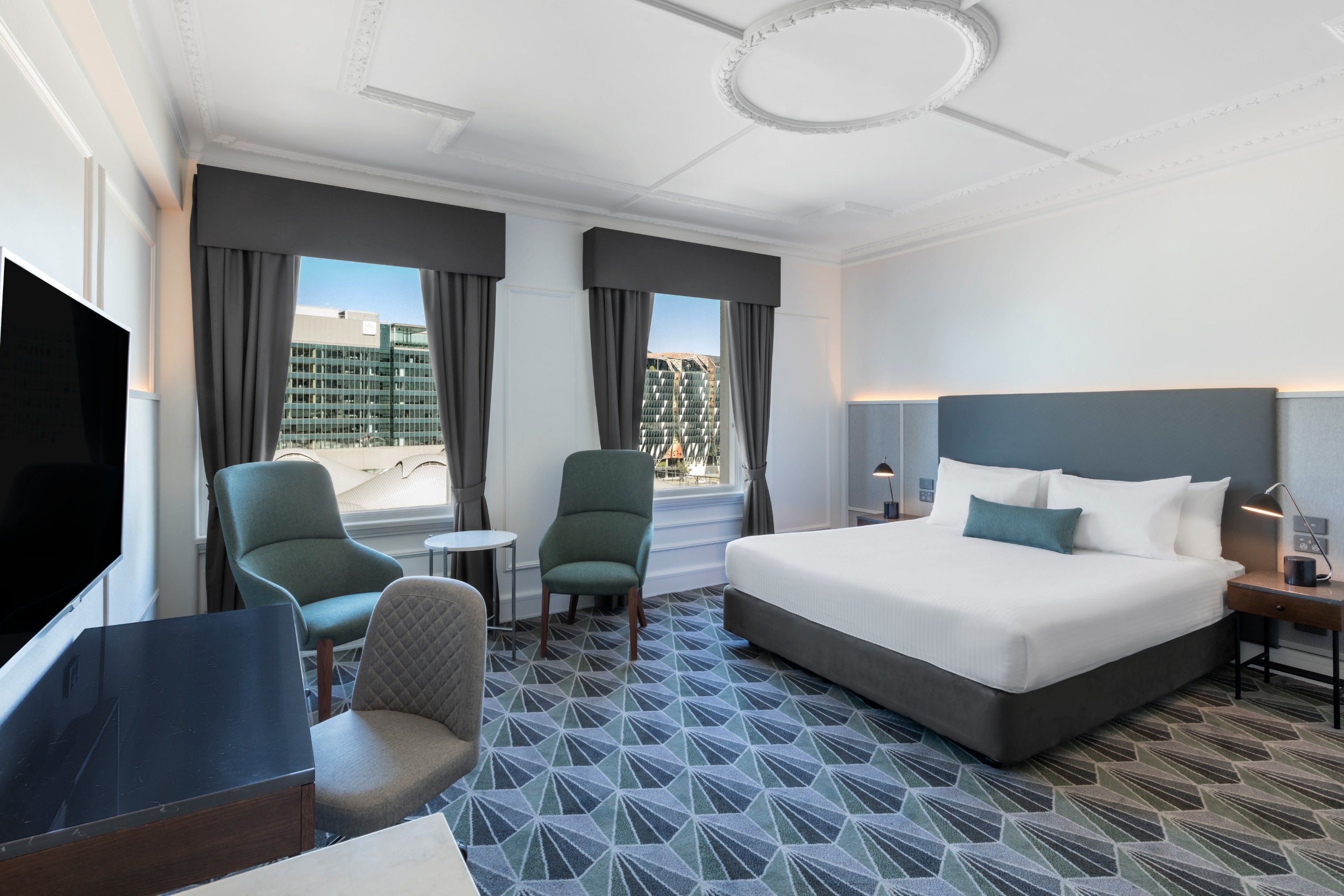 A hotel room with a large bed, two armchairs, a small table, a TV, and a view of a building through the windows.
