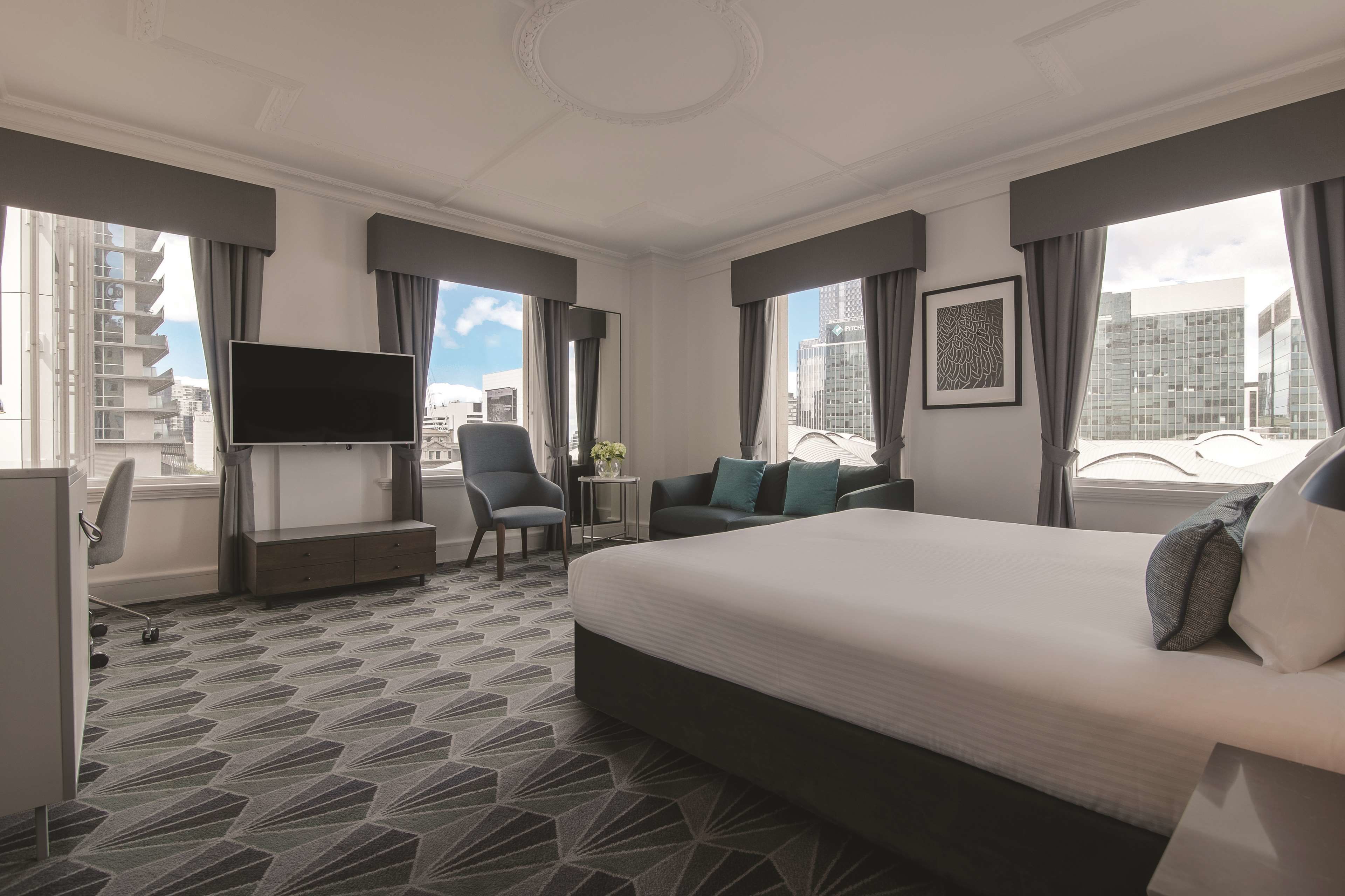 A hotel room with a large bed, a desk with a flat-screen TV, a chair, a sofa, and large windows with curtains.