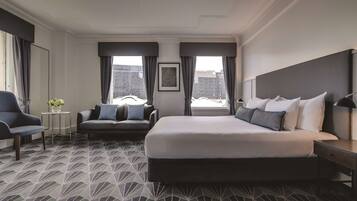 Habitación de hotel con una cama grande, un sofá, una silla, una mesita con un florero y un cuadro en la pared.