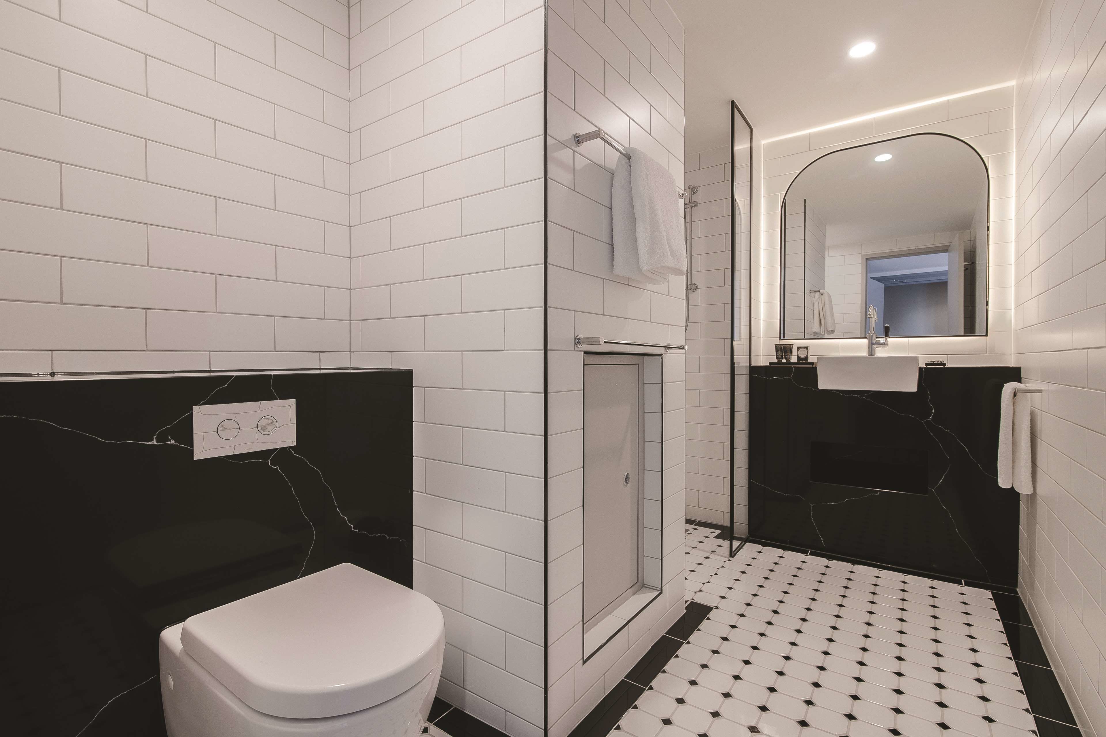Un baño moderno con azulejos blancos tipo metro, una pared de acento en mármol negro, un inodoro blanco, una ducha con cerramiento de vidrio y un suelo con estampado blanco y negro.