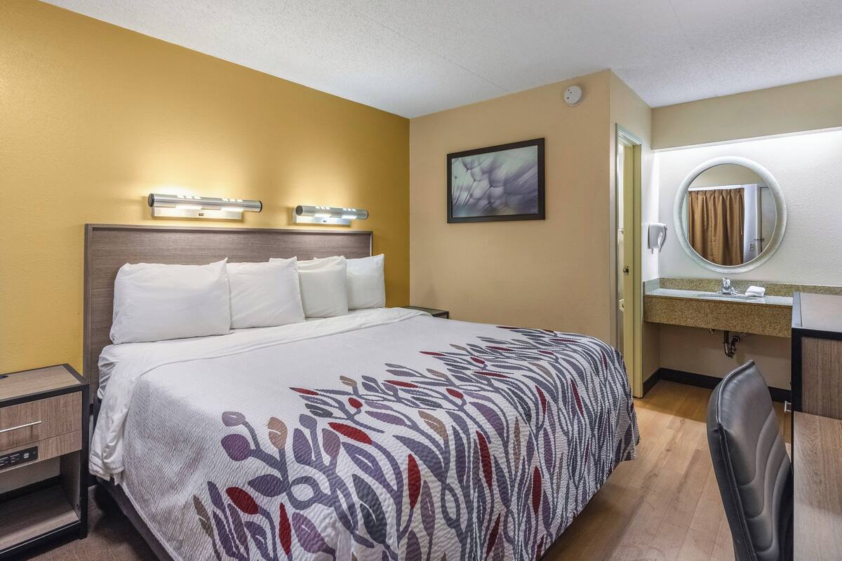 Ein Hotelzimmer mit Bett, Nachttisch und einem Badezimmer mit Waschbecken und Spiegel.