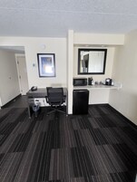 A hotel room with a desk, chair, trash bin, microwave, refrigerator, and a mirror on the wall.