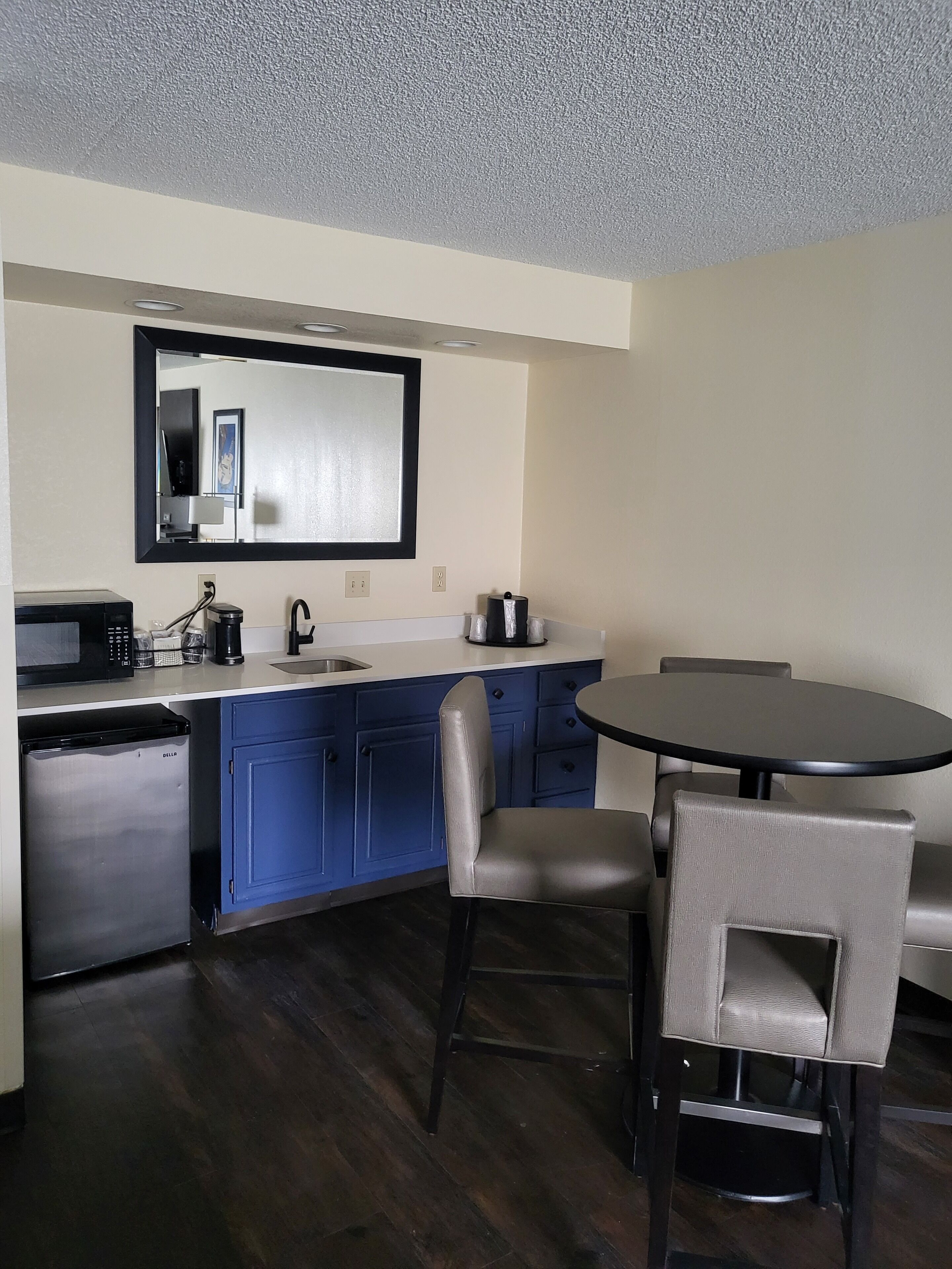 A compact kitchen with a microwave, coffee maker, and sink. There is a small refrigerator and a dining table with chairs.
