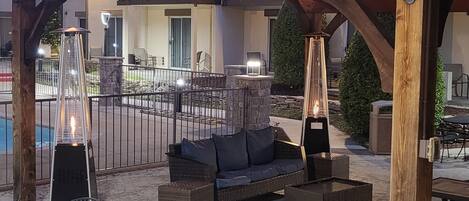 An outdoor patio with a pergola, seating area, and a pool in the background.