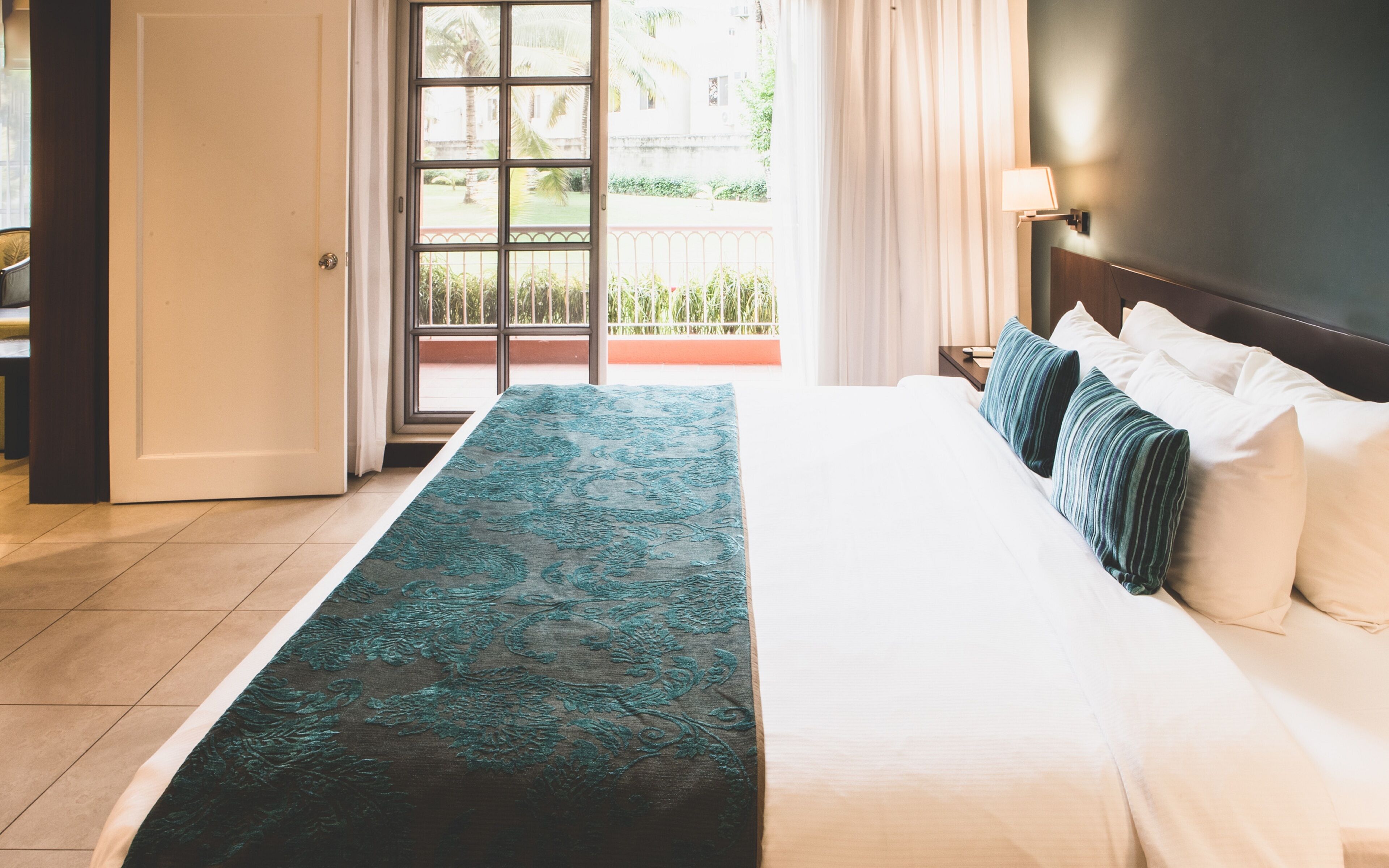 Una camera d'albergo con un letto, un comodino, una lampada e vista sull'esterno attraverso una finestra.