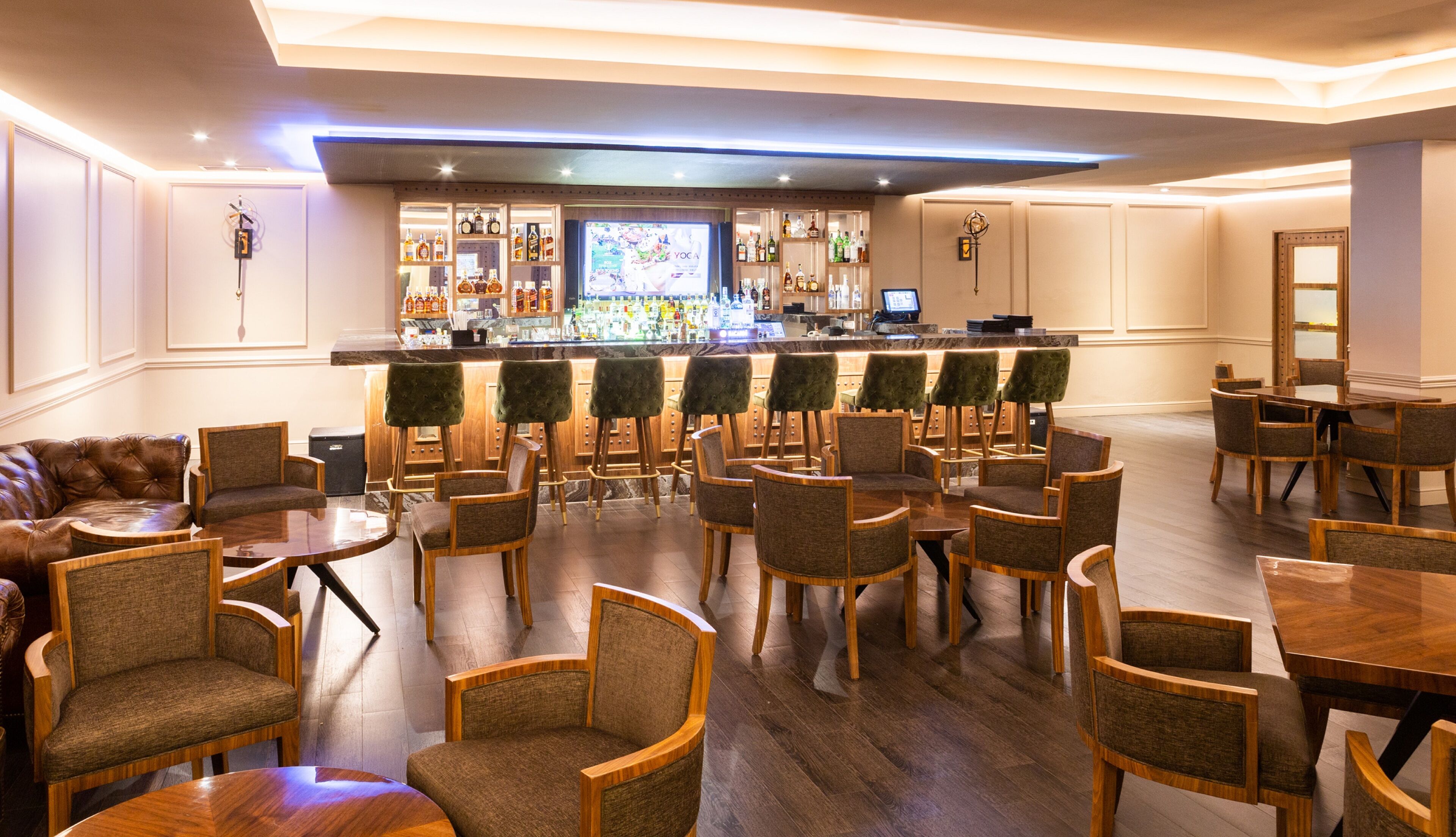 Uma área de bar com balcão, diversas opções de assento incluindo banquetas e cadeiras, e variedade de garrafas e copos em prateleiras.