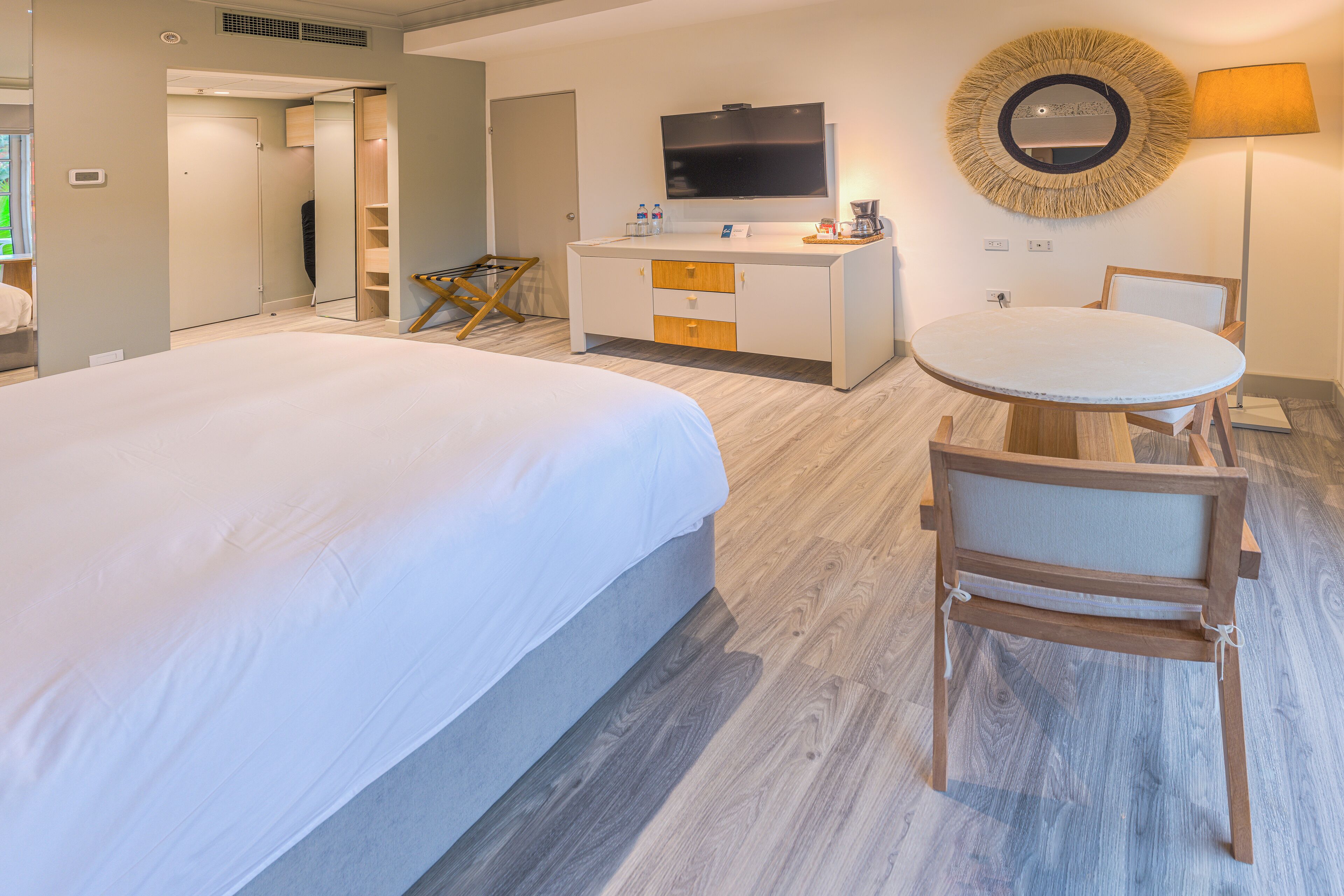 A hotel room with a large bed, a desk with a flat-screen TV, a round mirror, a small table with a chair, and a lamp.