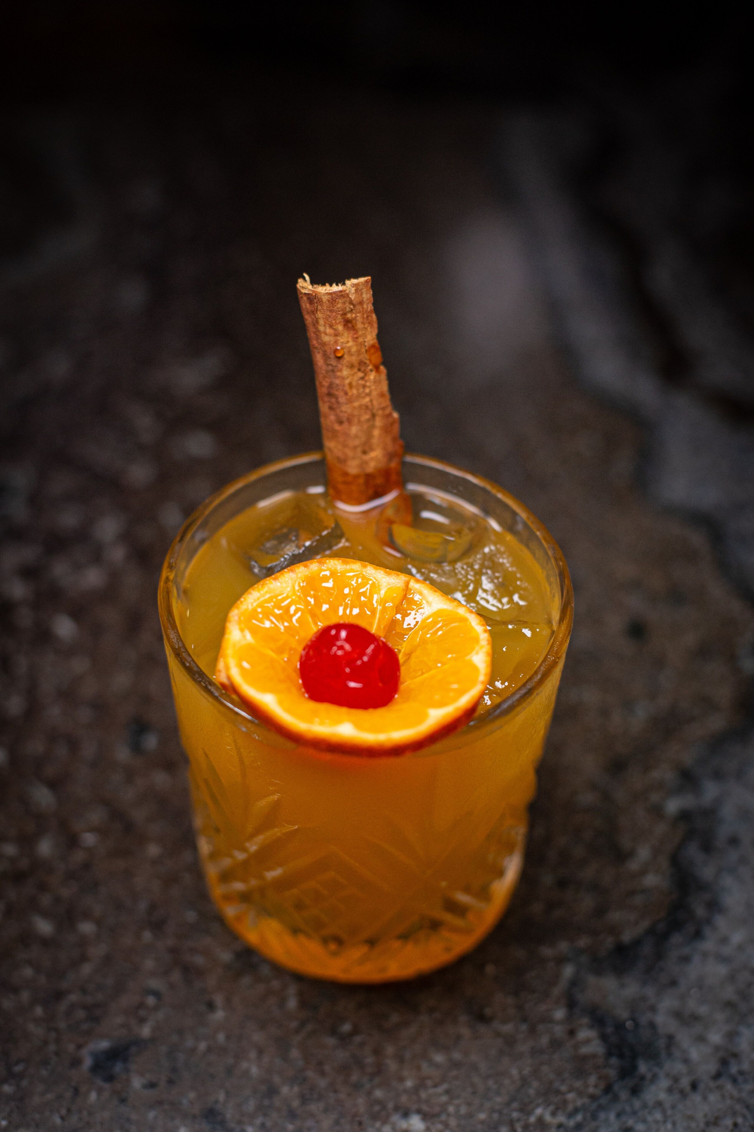 A glass of amber-colored beverage with a slice of orange and a cherry, garnished with a cinnamon stick, on a textured surface.